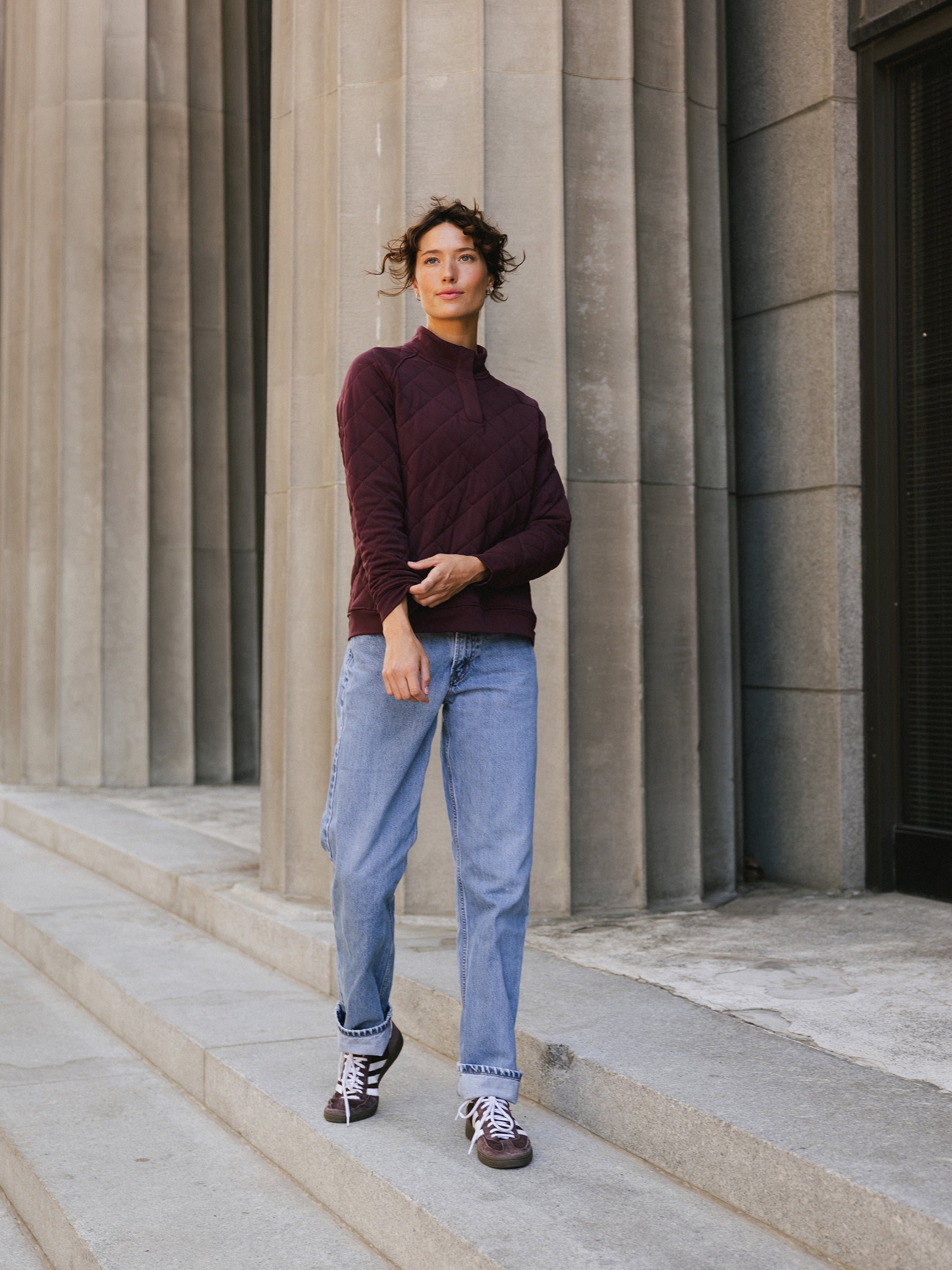 A person stands confidently on concrete steps in front of a building with tall columns. They are wearing a maroon Women's Ultra-Soft Bamboo Quilted Snap Pullover from Cozy Earth, light blue jeans, and maroon sneakers. The person has short, curly hair and appears relaxed with one hand holding their elbow. 