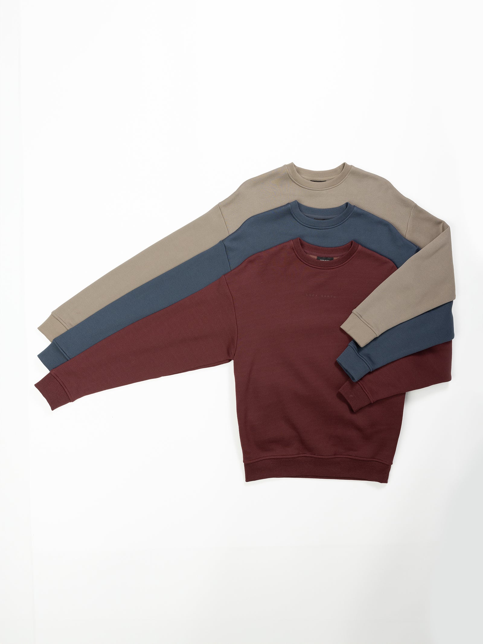 Three Women's CityScape Crewneck sweaters from Cozy Earth, available in beige, blue, and maroon, are neatly stacked on a plain white background. Each sweater features long sleeves and a simple design. 