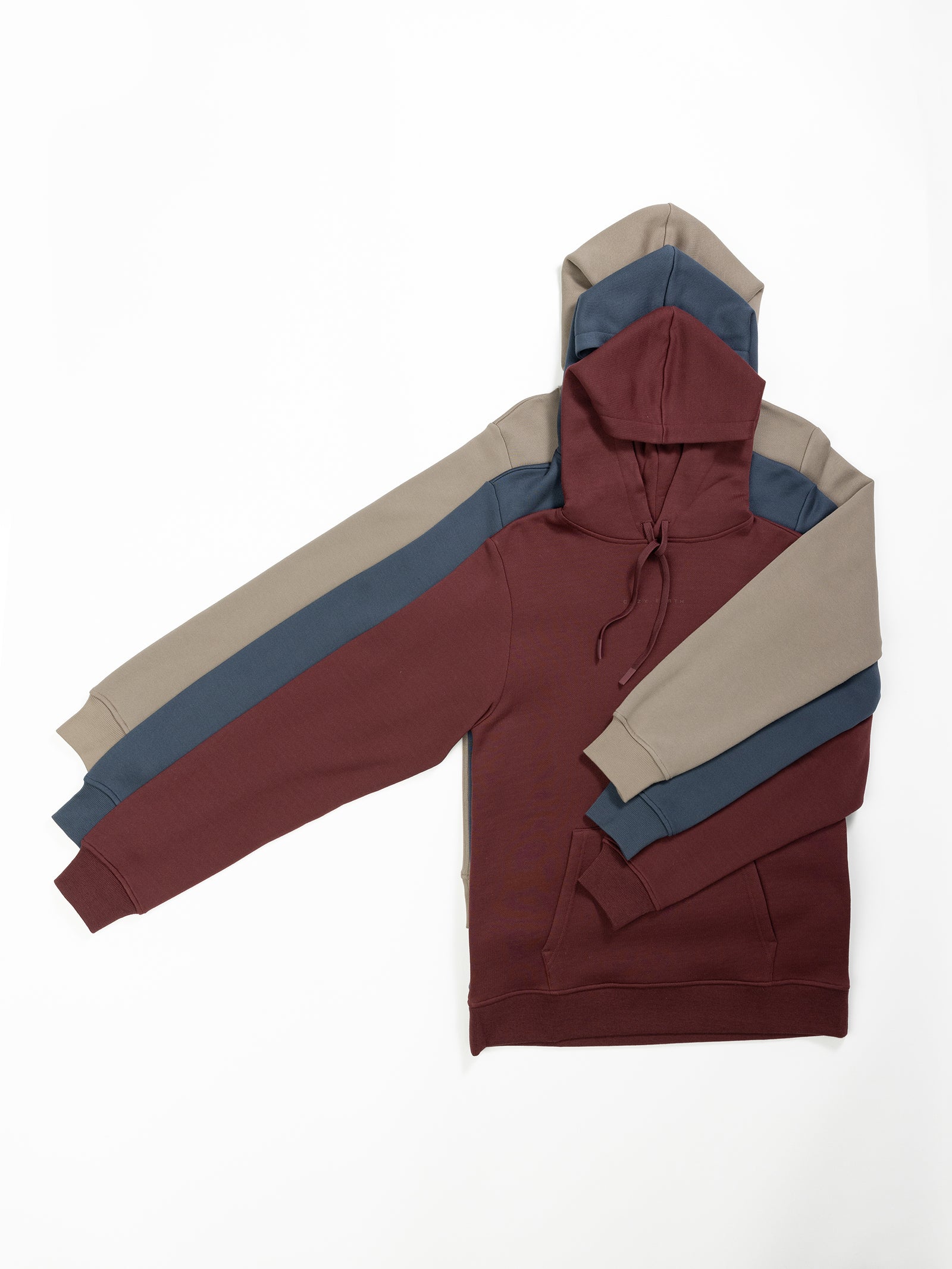 Three Men's CityScape Hoodies from Cozy Earth stacked on top of each other with their sleeves spread out. The bottom hoodie is maroon, the middle one is blue, and the top one is gray. All feature a front pocket and drawstrings for the hood. 