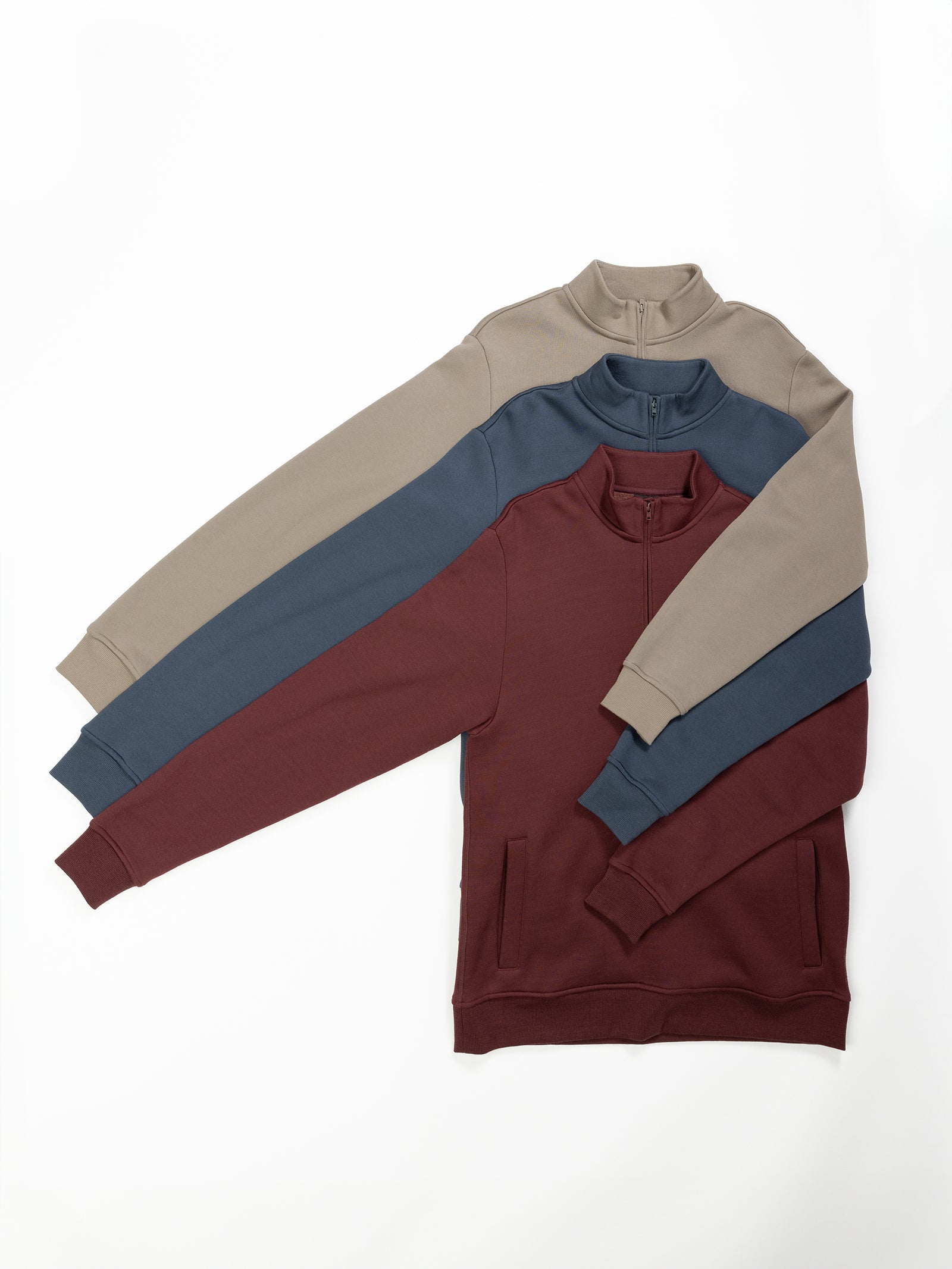 Three Men's CityScape Quarter Zip sweaters from Cozy Earth are laid on a white surface. The top sweater is beige with dark gray sleeves, the middle one is gray with teal sleeves, and the bottom one is maroon. They have a casual, comfortable appearance. 