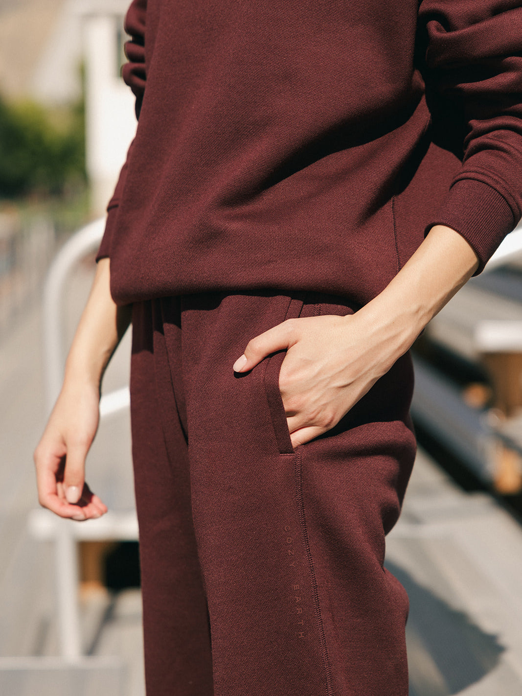 A person dressed in matching Cozy Earth Women's CityScape Sweatpants and a maroon sweatshirt stands outdoors with one hand in their pocket. Only the torso, right arm, and legs are visible. The background is blurred, suggesting an urban environment. 