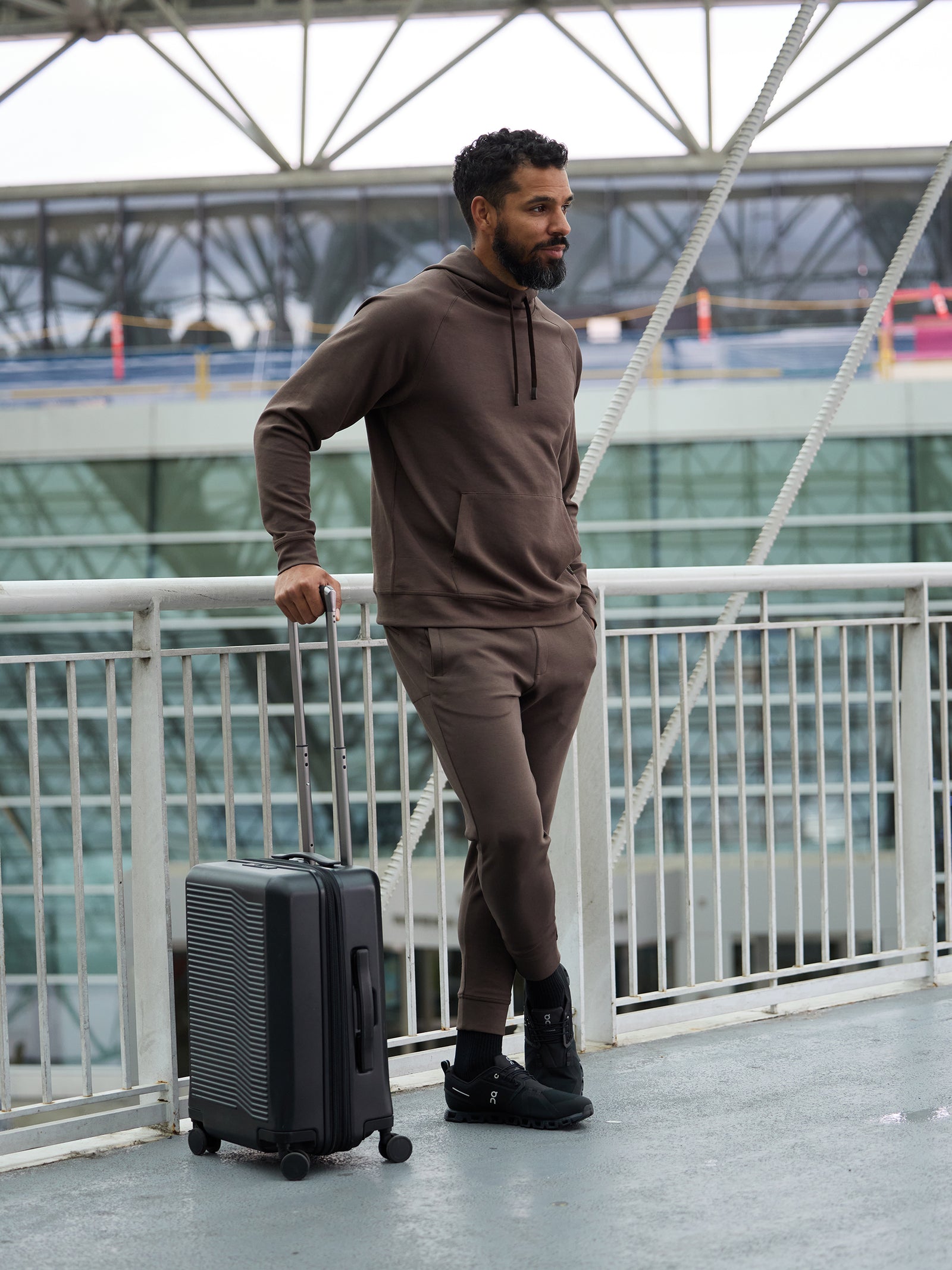 A bearded man wearing Cozy Earth's Men's StretchTech Jogger and a brown hoodie stands on a pedestrian walkway, leaning on a black roller suitcase. He looks to the side, with an airport terminal and palm trees visible in the background through the glass windows. 