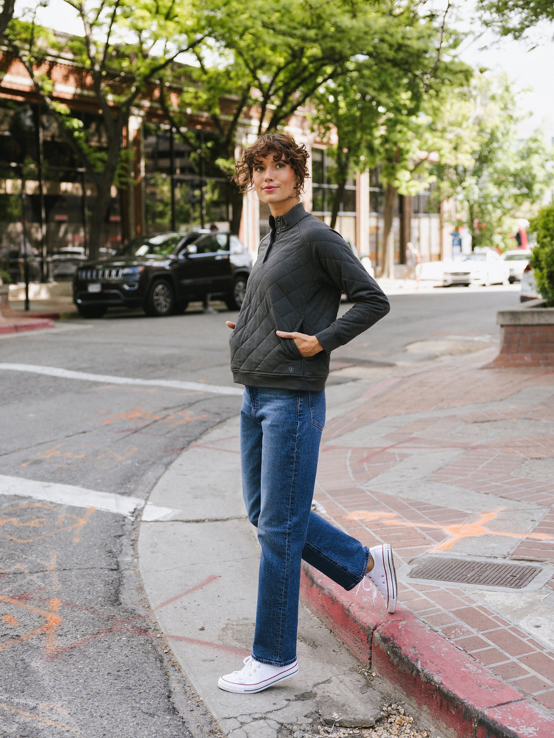 A woman with curly hair stands on a city street corner, wearing Cozy Earth's Women's Ultra-Soft Bamboo Quilted Snap Pullover, blue jeans, and white sneakers. She has her hands in her pullover pockets and is leaning against a brick ledge with a sidewalk and trees in the background. 