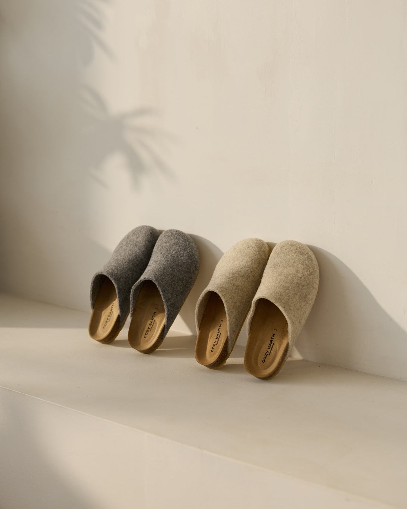 Two pairs of Cozy Earth's Lakehouse Clog slippers, one gray and one beige, with wooden soles and a minimalist design, are placed side by side on a light surface. A soft shadow of a plant appears on the wall. 