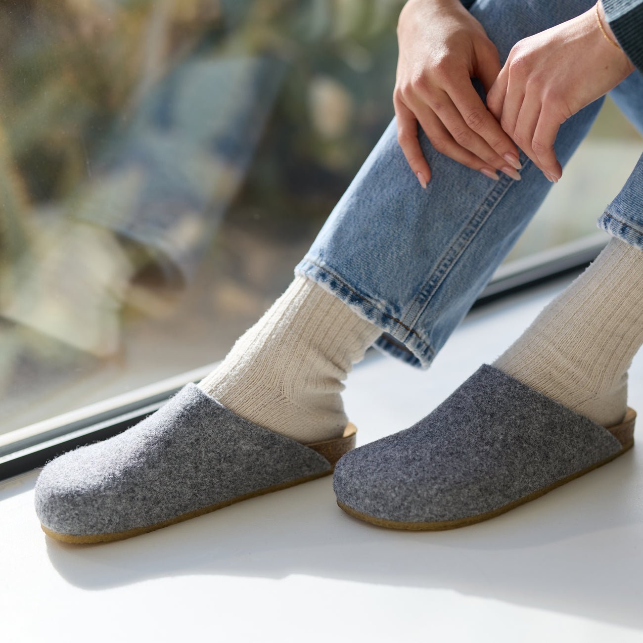 A person in light blue jeans and cream socks is seated near a window, showing their lower legs and hands while wearing Cozy Earth Lakehouse Clogs with gray woolen uppers and cork soles. The light creates a soft shadow on the surface. 