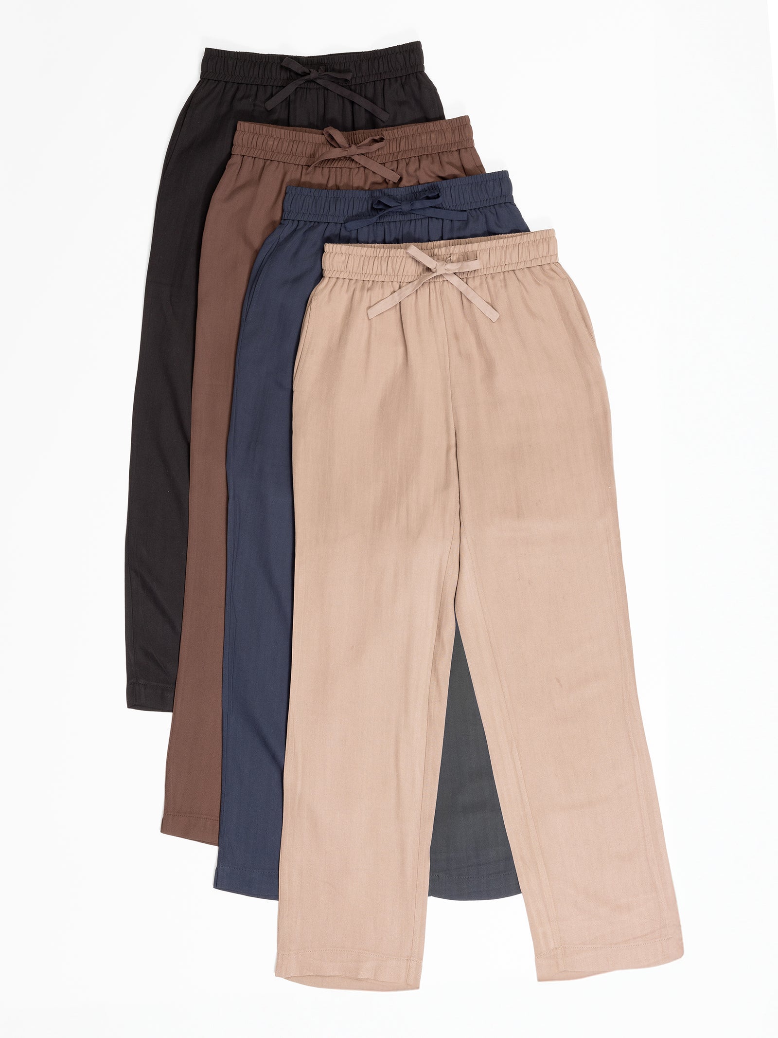 Four pairs of the Women’s Sunset Cropped Pant by Cozy Earth are laid flat in a fan arrangement. The pants come in various colors: black, brown, navy blue, and beige. Each pair features an elastic waistband with a drawstring. The fabric appears soft and lightweight. 