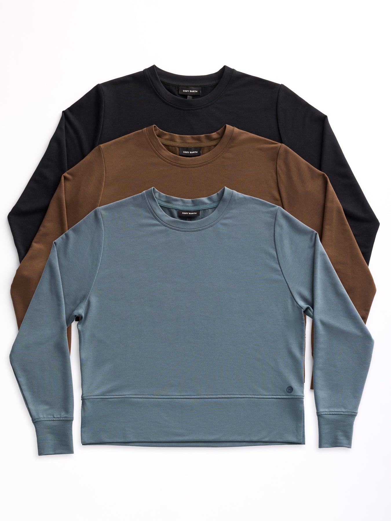 Three Women's StudioLite Crewneck shirts from Cozy Earth, in black, brown, and blue, are stacked on a flat surface in descending order. They feature simple solid colors without visible patterns or logos. 