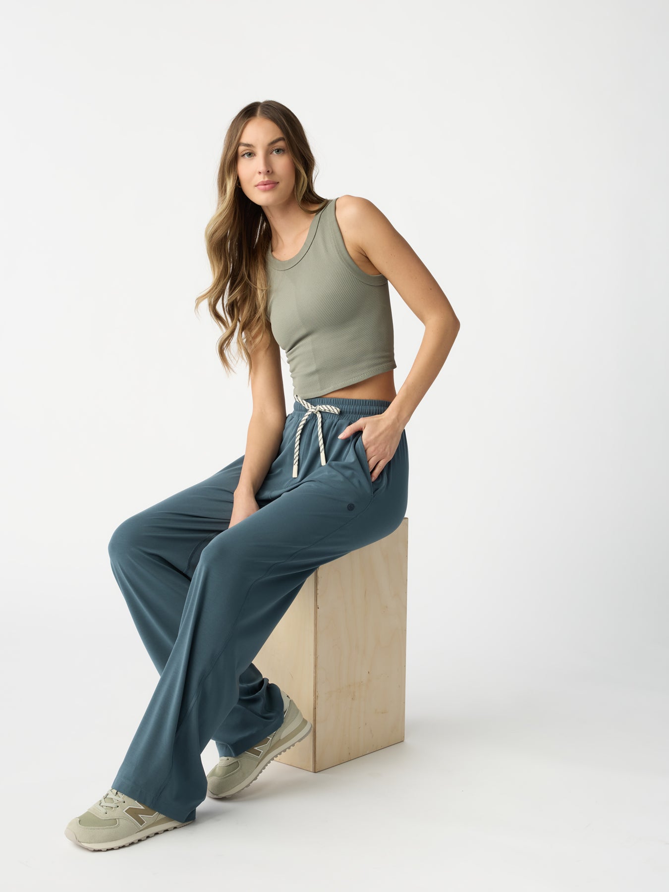 A person with long hair sits on a wooden box, wearing a light green sleeveless top, Cozy Earth's Women's Studio Wide Leg Pants in blue featuring a drawstring, and beige sneakers against a plain white background. 