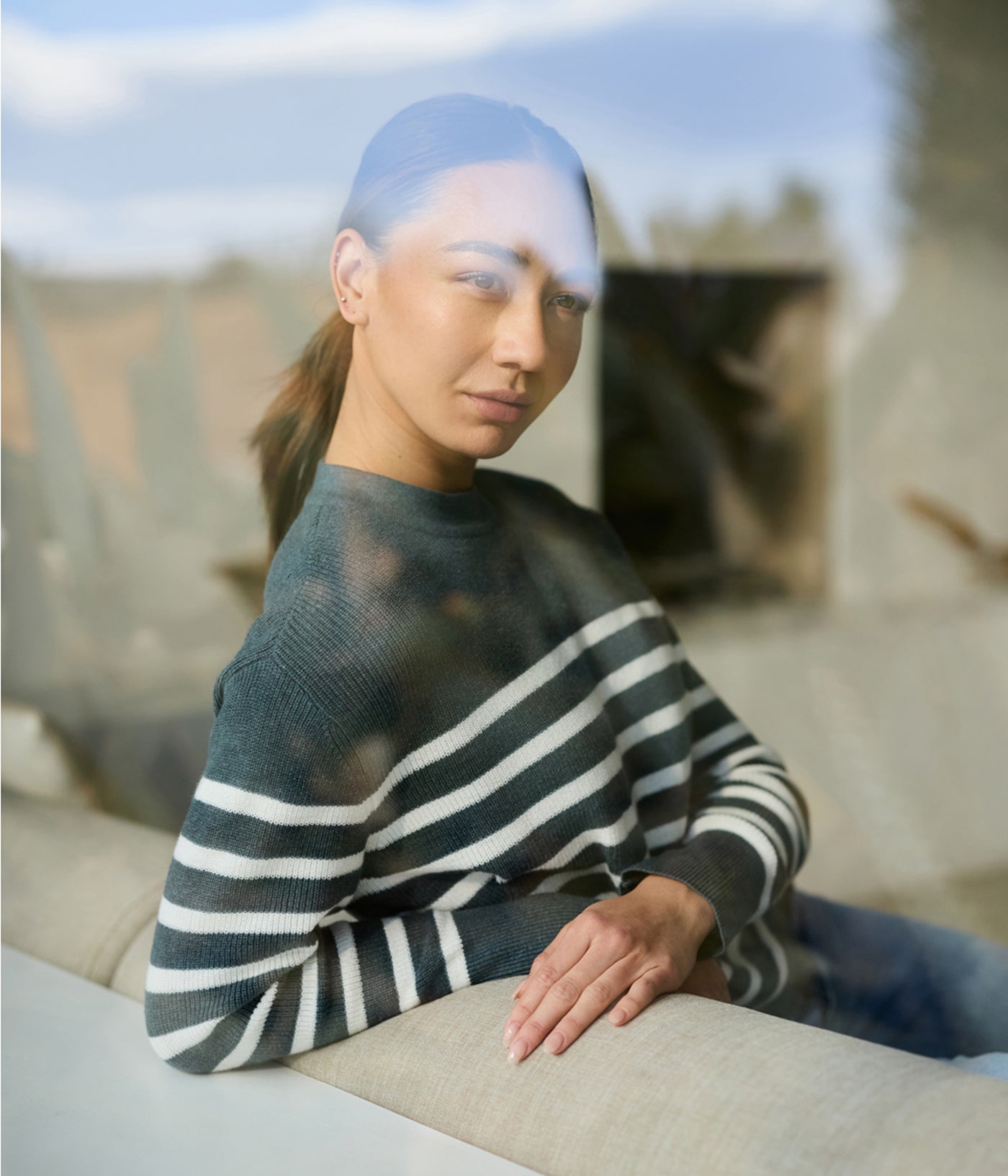 A woman with a ponytail gazes out a glass window indoors, wearing the striped Women's Rowan Sweater by Cozy Earth. The glass reflection blurs the outdoor scenery. 