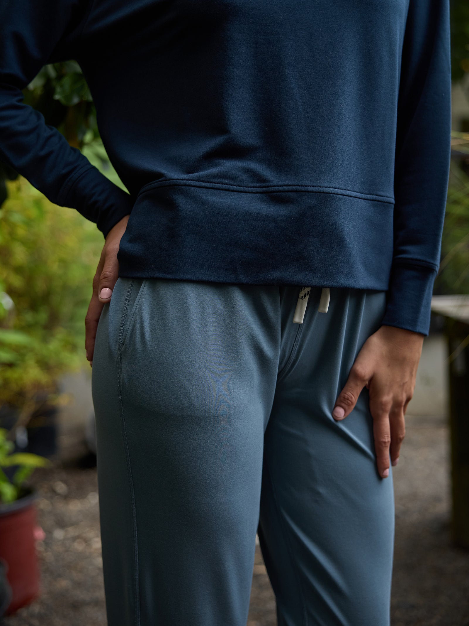 A person dressed in a dark blue long-sleeve shirt and light blue Women's Studio Jogger from Cozy Earth stands outdoors, with one hand on their hip and the other resting on their thigh. The background features blurred greenery and potted plants. 