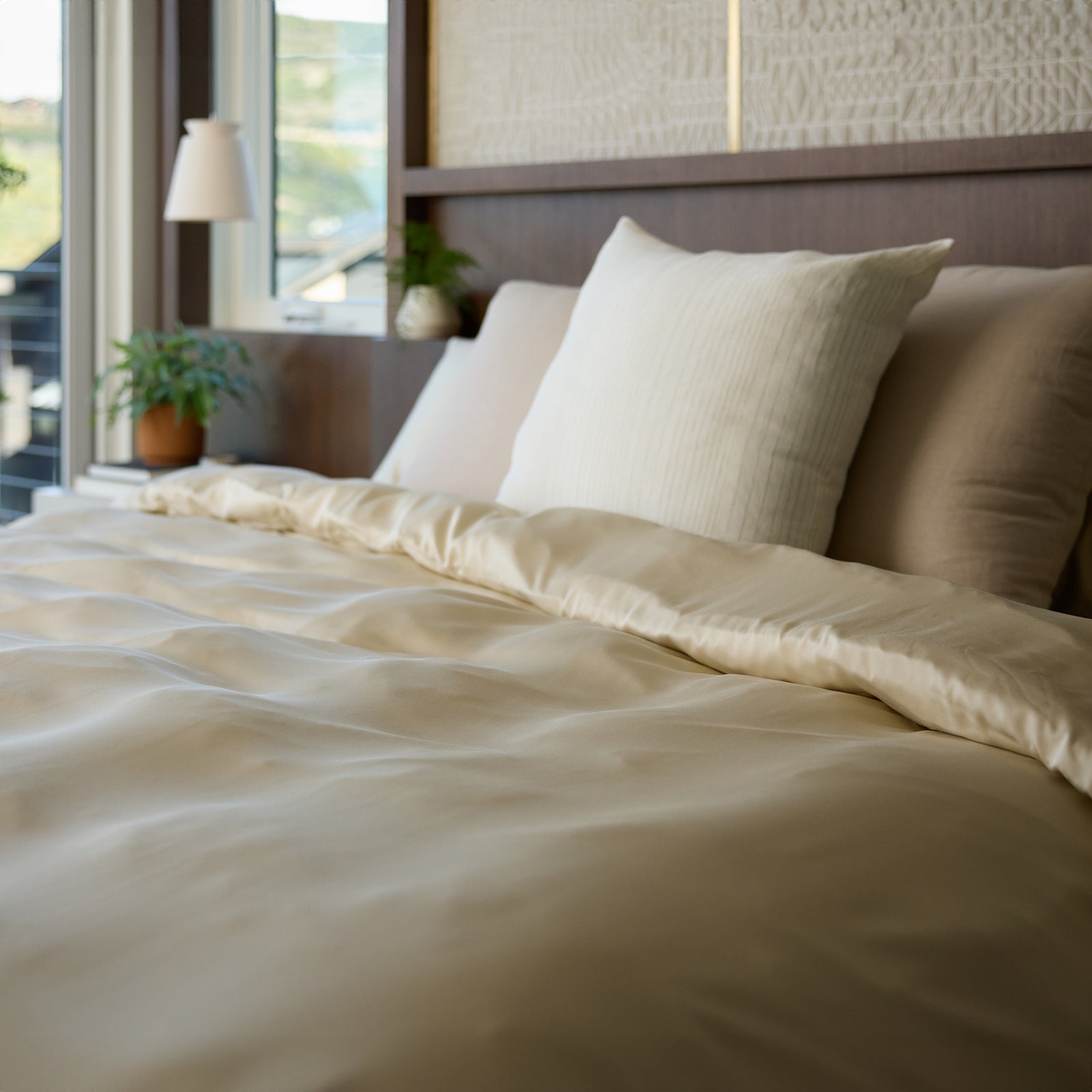 A neatly made bed with a creamy, soft Down Alternative Comforter by Cozy Earth and several pillows. Natural light enters through a window, illuminating a plant on the bedside table. The ambiance is serene and cozy.