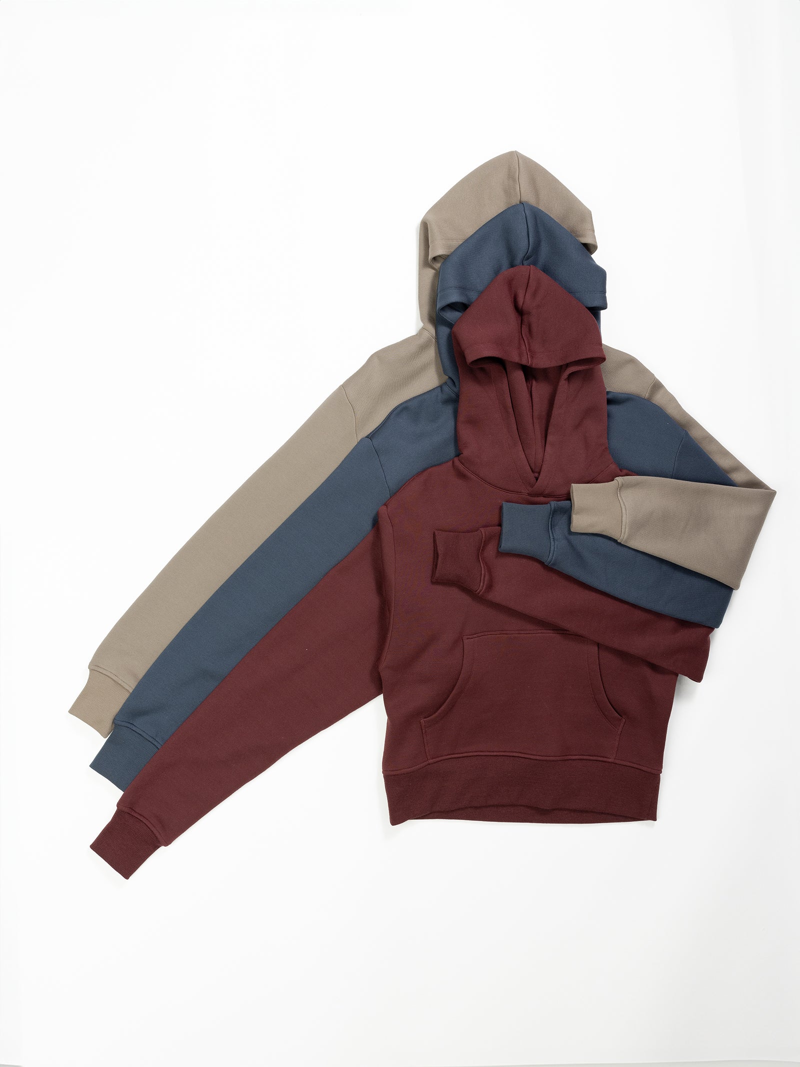 Four Women's CityScape Cropped Hoodies by Cozy Earth are neatly arranged overlapping each other on a plain white background. The colors from top to bottom are beige, navy blue, dark teal, and maroon. Each hoodie features a front pocket and offers a relaxed fit. 