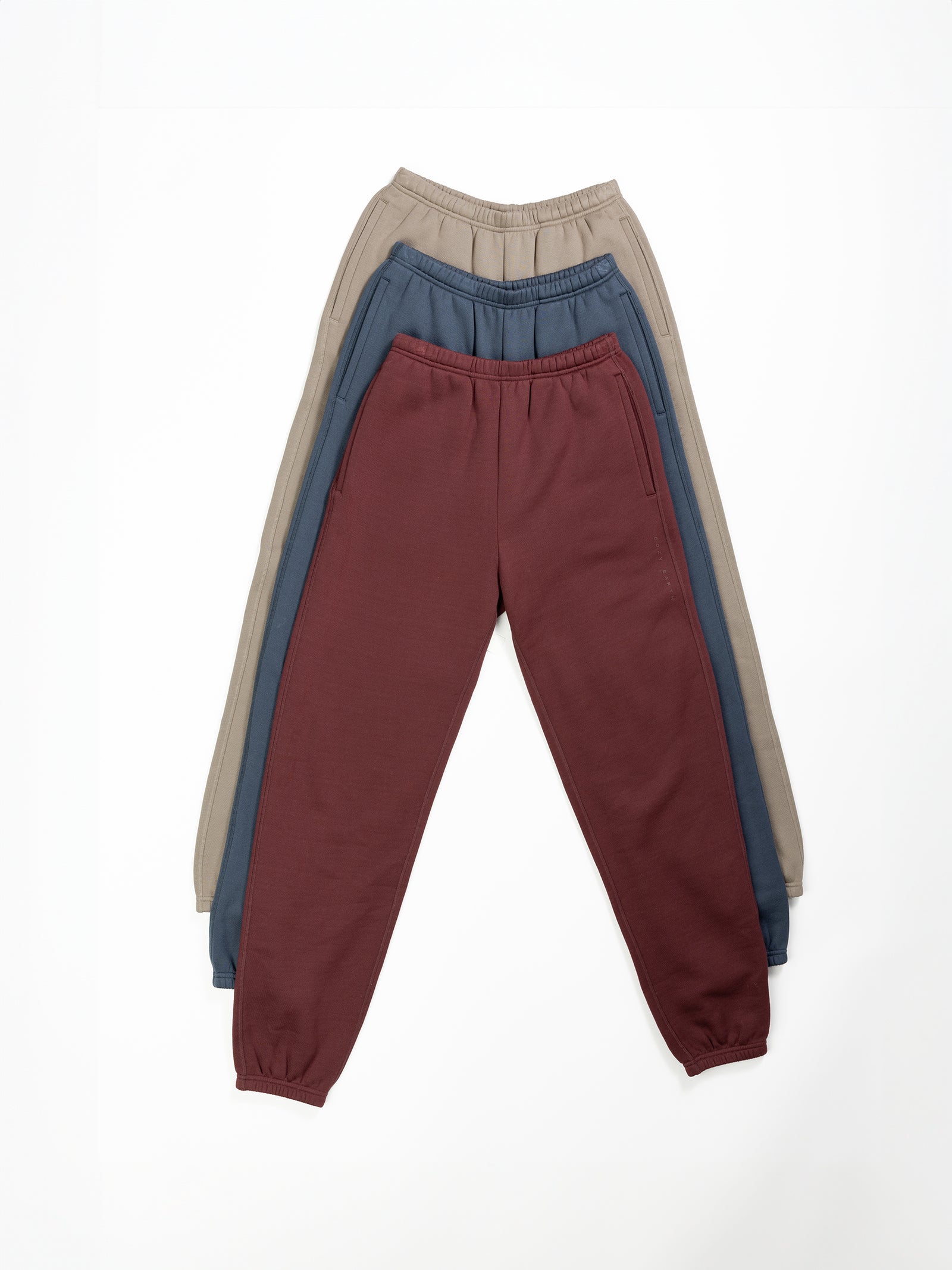 Three pairs of Women's CityScape Sweatpants from Cozy Earth arranged in a stack, partially overlapping each other. The sweatpants on top are maroon, the middle pair is dark blue, and the bottom pair is beige. All of them feature an elastic waistband and cuffs at the ankles. 