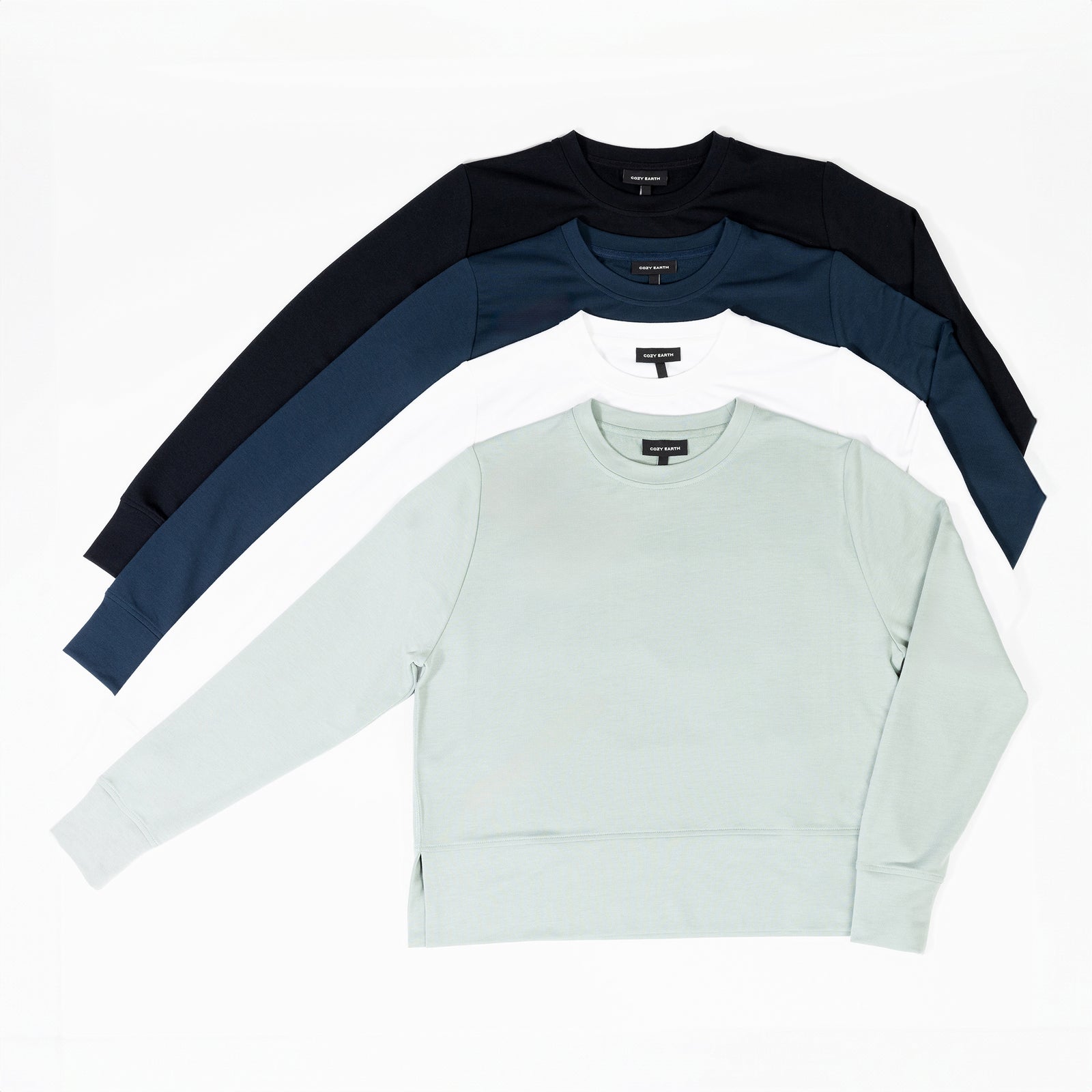 Three Women's StudioLite Crewnecks from Cozy Earth are laid out in a fan arrangement on a flat surface. From top to bottom, the colors are dark navy blue with black sleeves, white, and light mint green. All have long sleeves and feature a simple, minimalist design. 