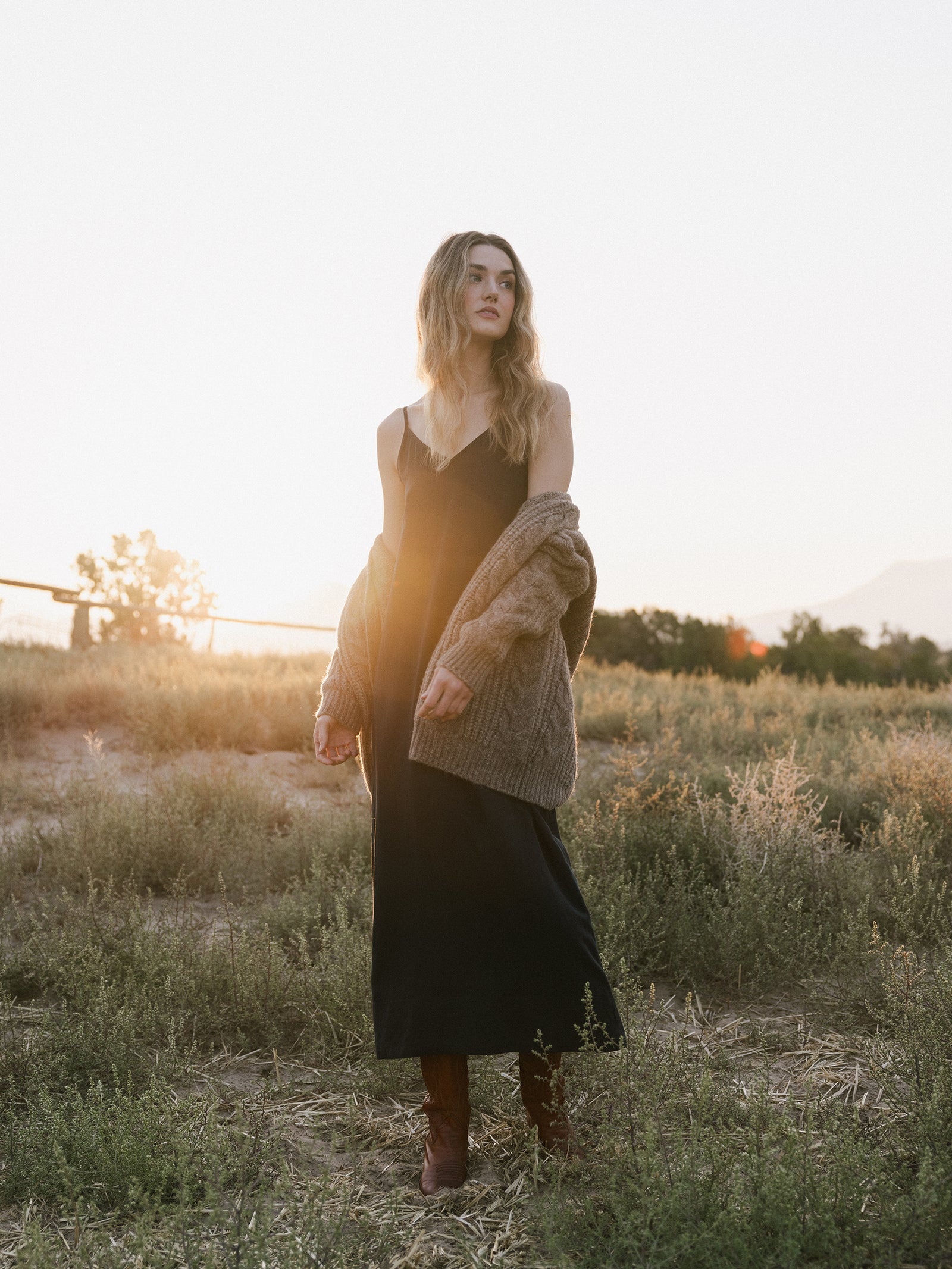 A woman with long wavy hair stands in a field at sunset. She is wearing Cozy Earth's Women's Sunset Slip Dress, a cozy brown cardigan draped over her shoulders, and brown boots. She gazes off into the distance as the sun casts a warm glow around her. 