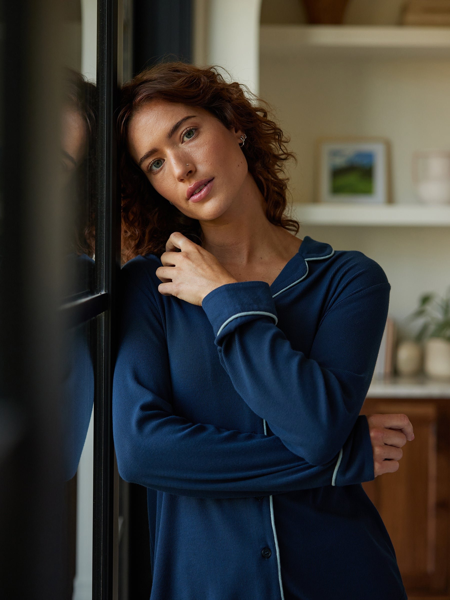 A person with curly hair, wearing a Cozy Earth Women's Bamboo Rib Knit Classic Long Sleeve Pajama Top in blue, stands thoughtfully by a window with one hand resting on their opposite shoulder. The background features shelves adorned with framed pictures and potted plants. 
