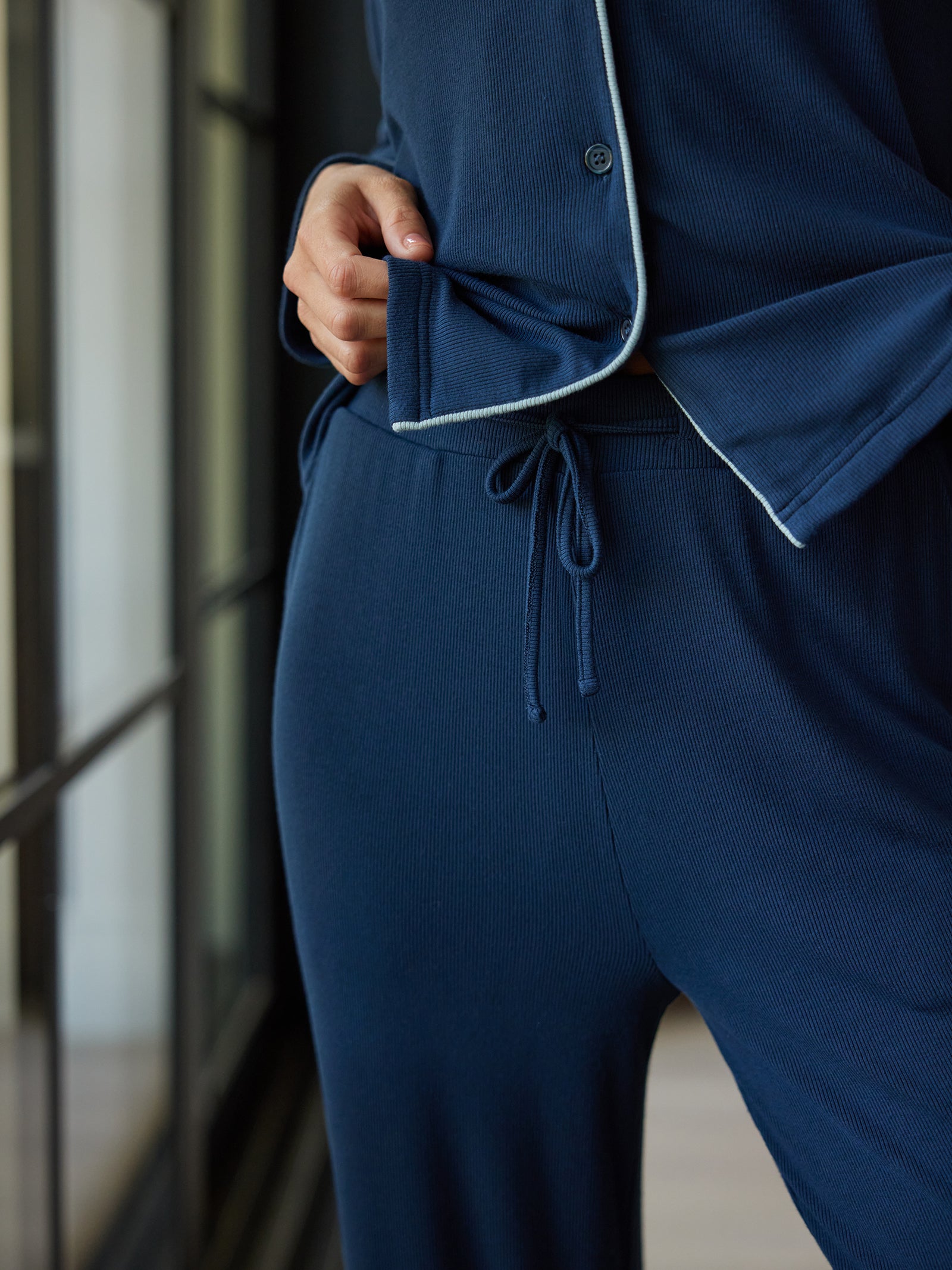 Close-up of a person wearing Cozy Earth's Women's Bamboo Rib Knit Classic Pajama Pant in navy blue. The photo focuses on their hand tucking into one of the pajama top pockets and the drawstring waistband of the pajama pants. The fabric has a ribbed texture, and the hem is contrasted with white piping. 