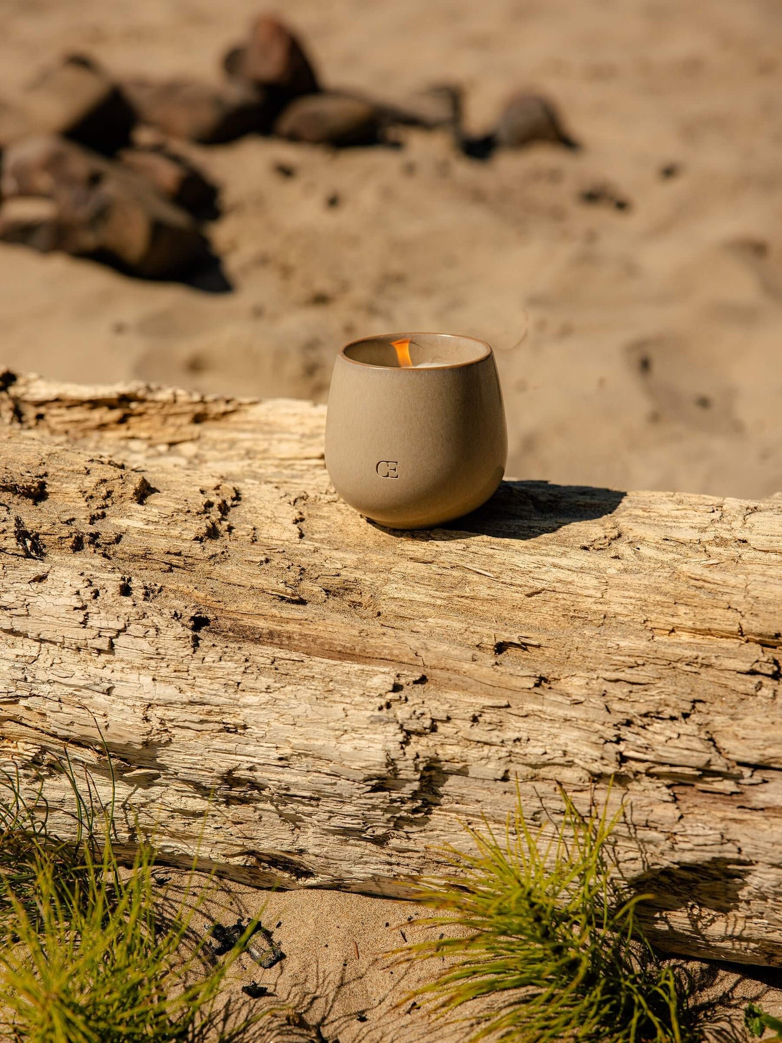 The Candle EMBER by Cozy Earth, in beige ceramic, rests on a weathered log on a sandy beach with rocks and beach grass in the background.