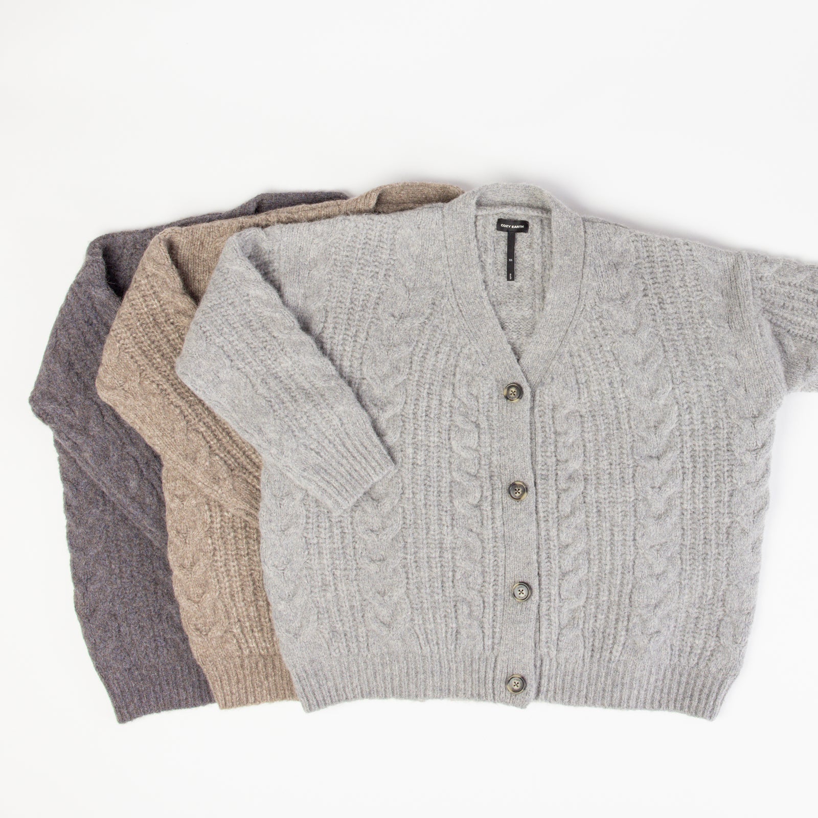 Three Oversized Cable Knit Cardigans by Cozy Earth, in gray, beige, and dark gray, are neatly folded and stacked on a white background. Each cardigan features a V-neck design and button closure. 