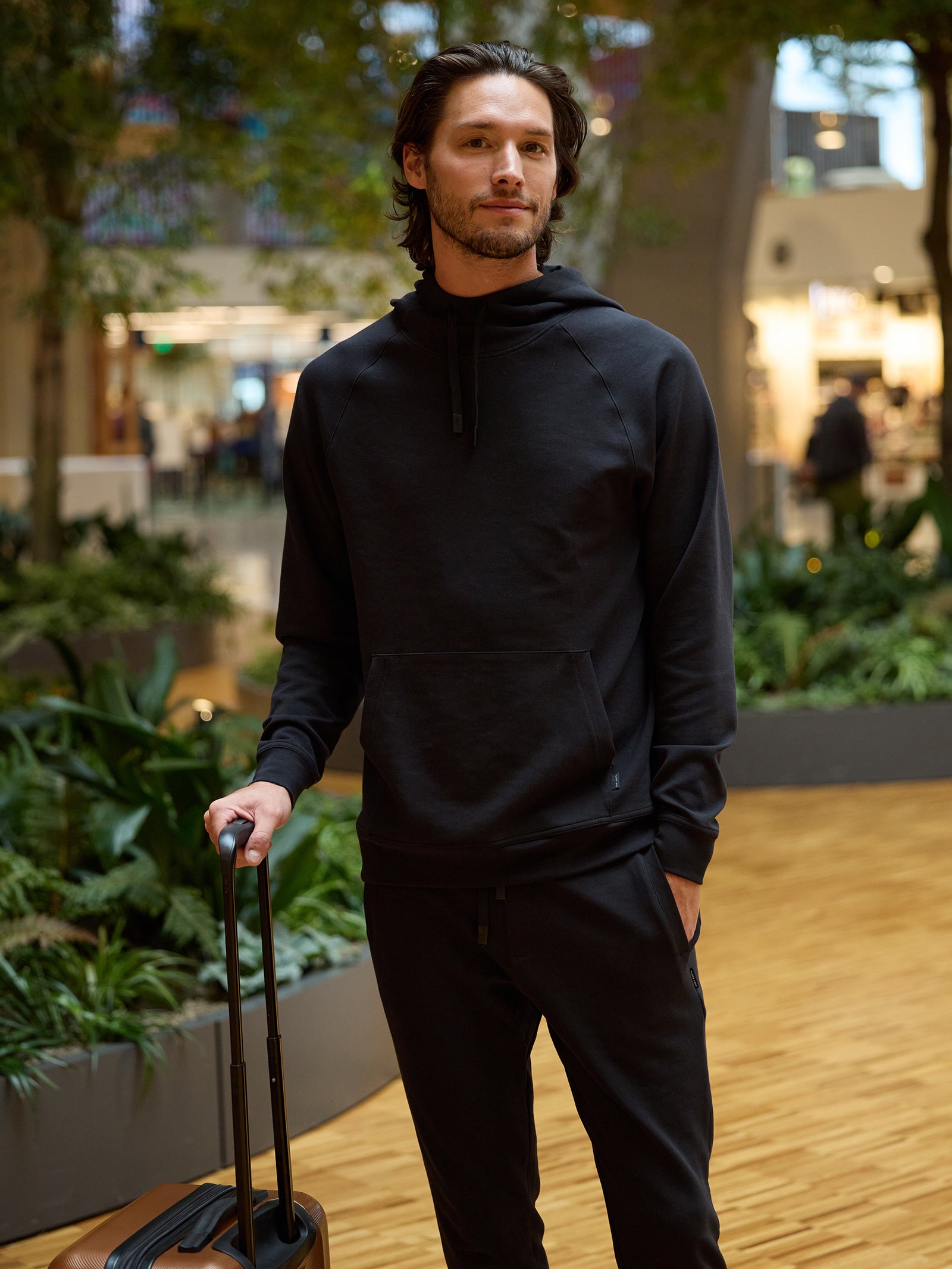 A man stands in a modern indoor setting with greenery and shops in the background. He wears a black Cozy Earth Men's StretchTech Hoodie and matching sweatpants, holding the handle of a brown suitcase. He has medium-length dark hair and a relaxed expression. 