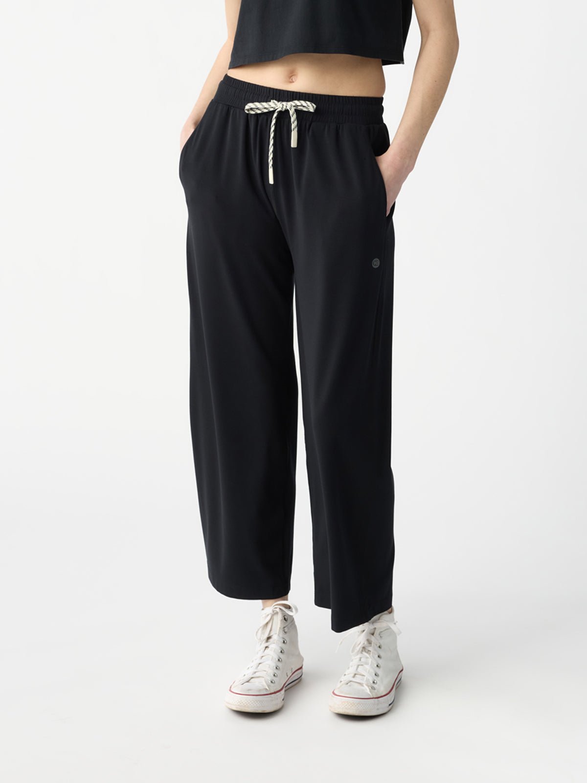 A person in Cozy Earth's Women's Studio Cropped Wide Leg Pant and a black cropped top stands against a plain background. The loose-fitting black pants, paired with white high-top sneakers, have their hands comfortably placed in the pockets. |Color:Jet Black