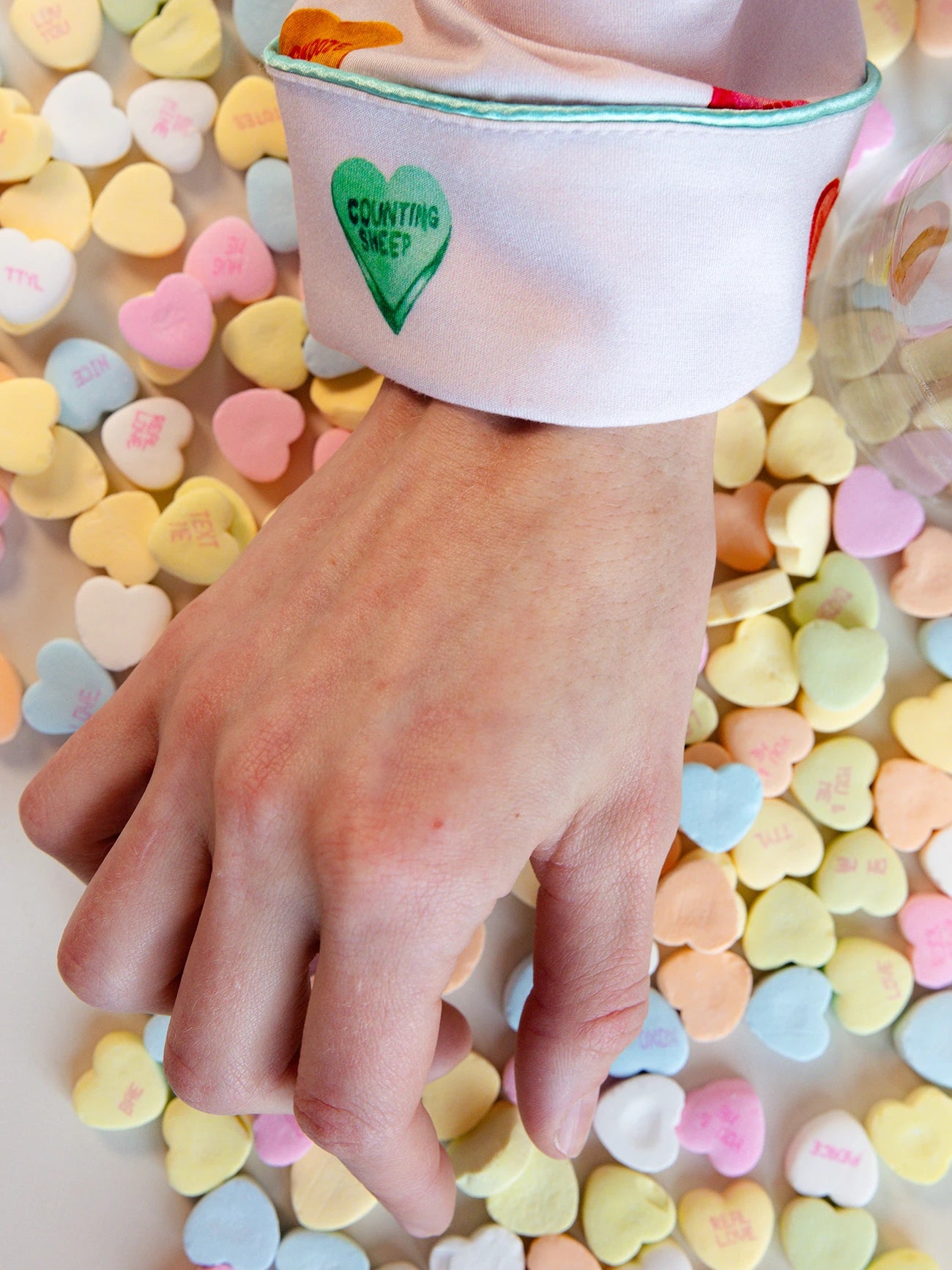 A hand with a Women's Long Sleeve Bamboo Pajama Top in Stretch-Knit by Cozy Earth, rests on a table covered with colorful candy hearts. 