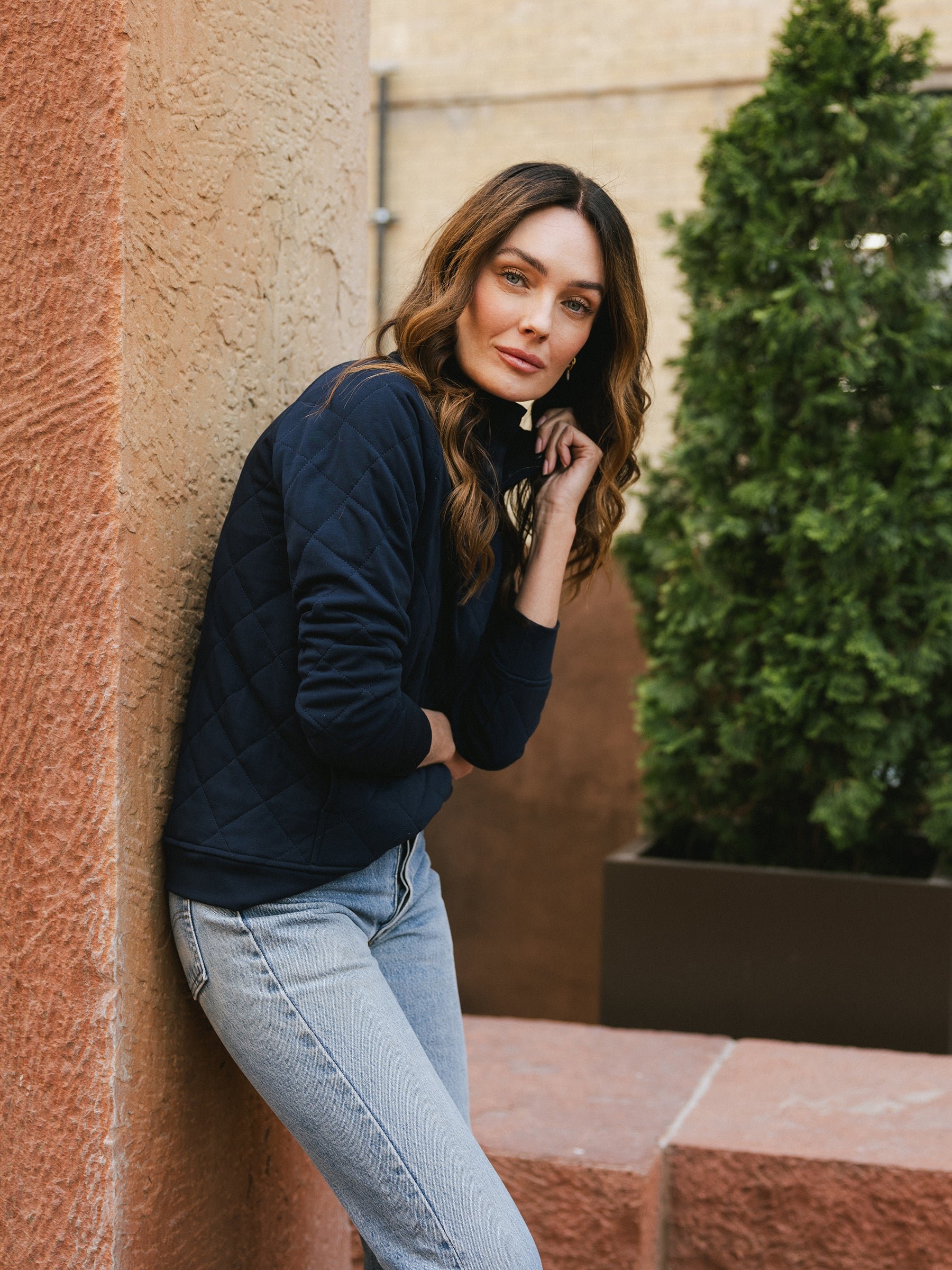 A person with long, wavy hair wearing a dark Women's Ultra-Soft Bamboo Quilted Snap Pullover from Cozy Earth and light blue jeans is leaning against a textured wall. They have a relaxed expression and their hand is touching their chin. In the background, there is a green potted plant. 