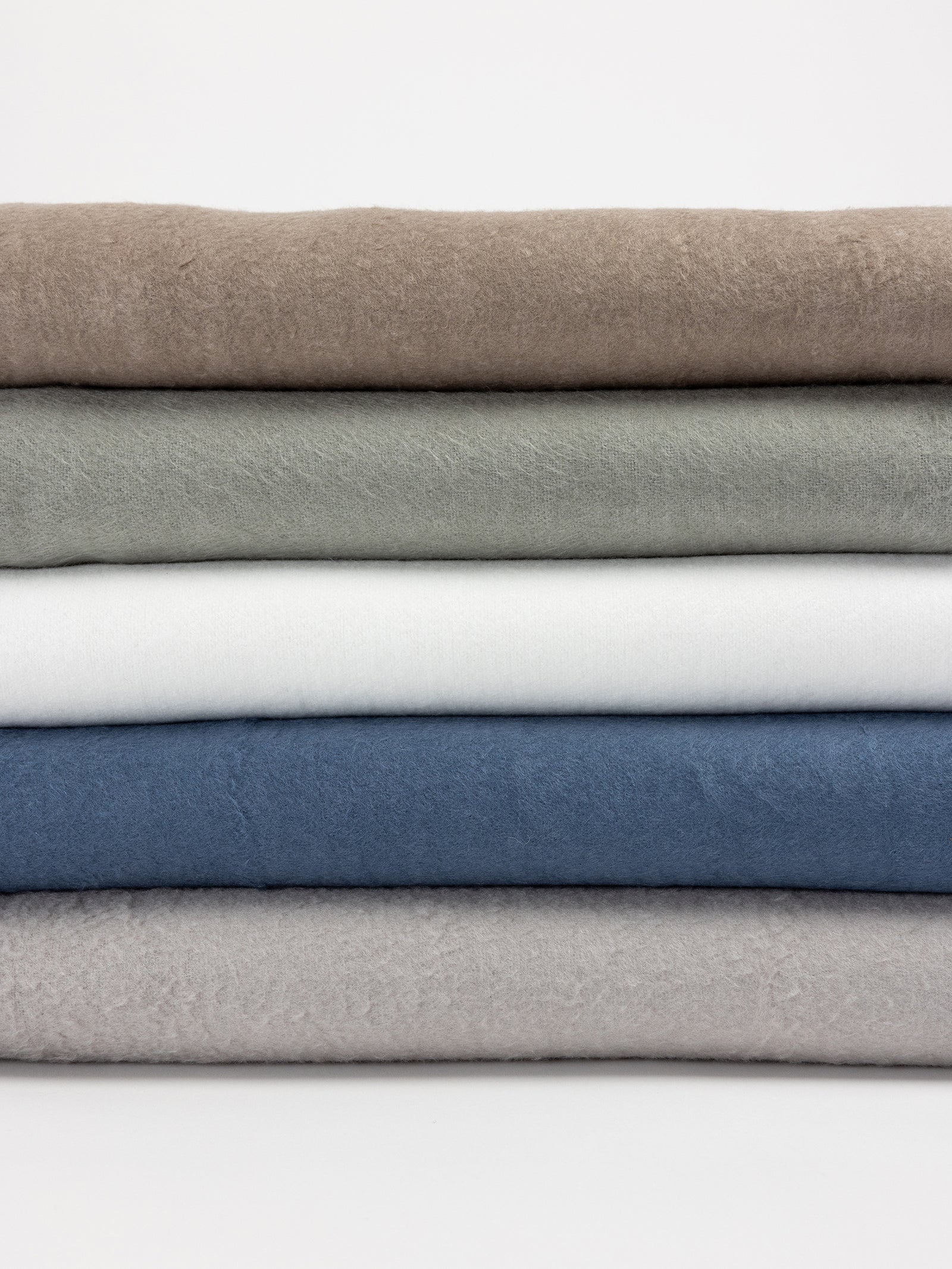 A neatly folded stack of five Bamboo Blankets by Cozy Earth, featuring a variety of colors. From top to bottom, the blankets are light brown, sage green, white, blue, and light gray. The background is white. 