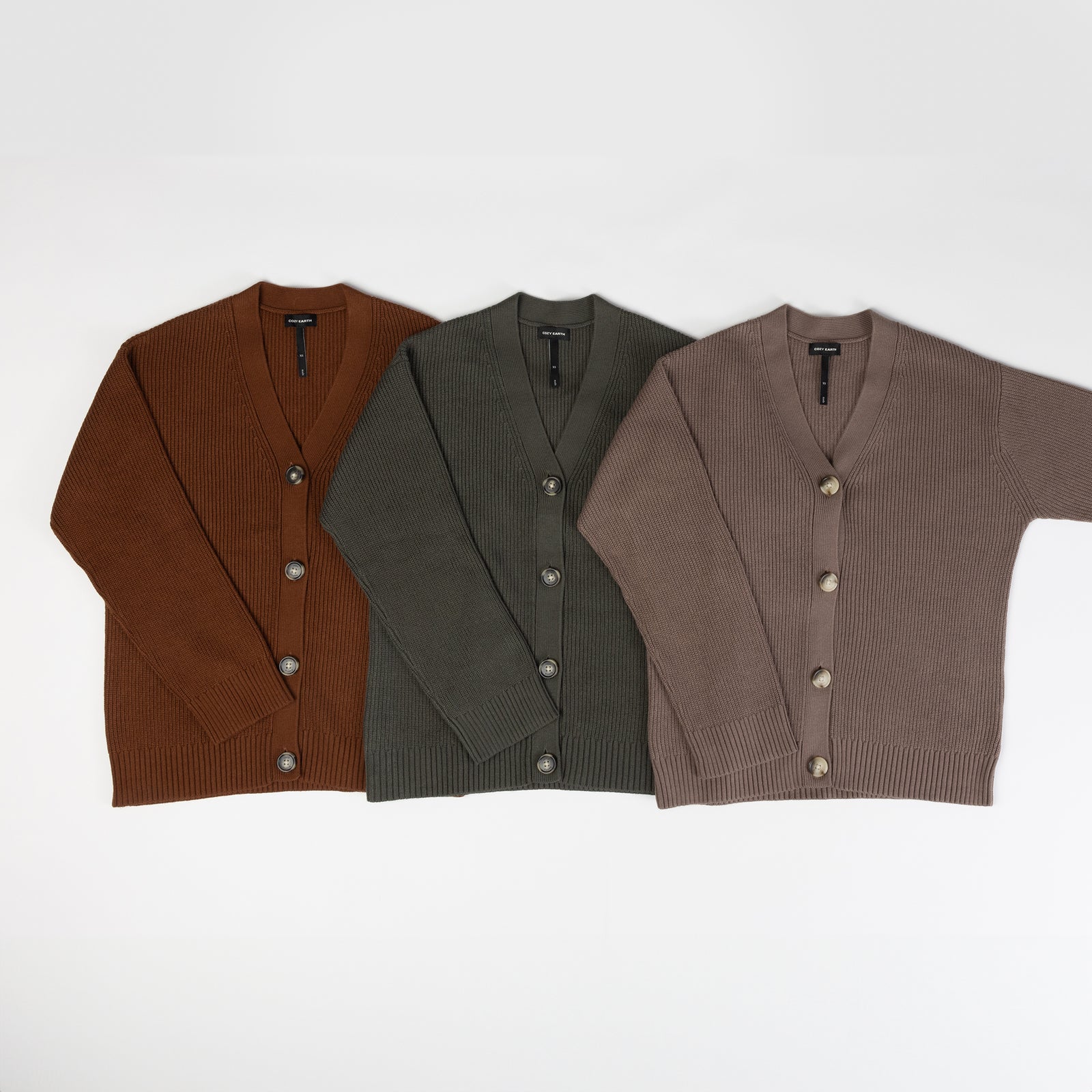 Three "Oversized Classic Cardigans" from Cozy Earth are laid out side by side. From left to right, they are in shades of brown, dark green, and taupe, each featuring a V-neckline with buttons running down the front against a plain white background. 