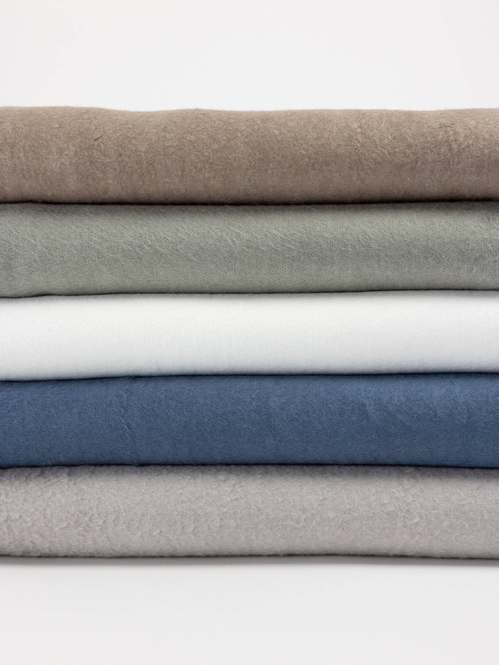 A stack of five folded Bamboo Blankets by Cozy Earth are arranged from top to bottom in beige, light olive, white, blue, and light gray. The plain white background highlights the texture and colors of these solid pieces. 