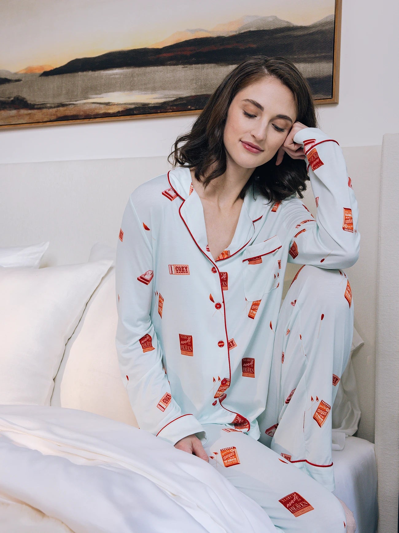 A woman with long brown hair sits on a bed in Cozy Earth Women's Stretch-Knit Bamboo Pajama Pant, light blue with red accents and match book patterns. Her eyes are closed, resting her head on her hand. A framed landscape painting is on the wall behind her. 