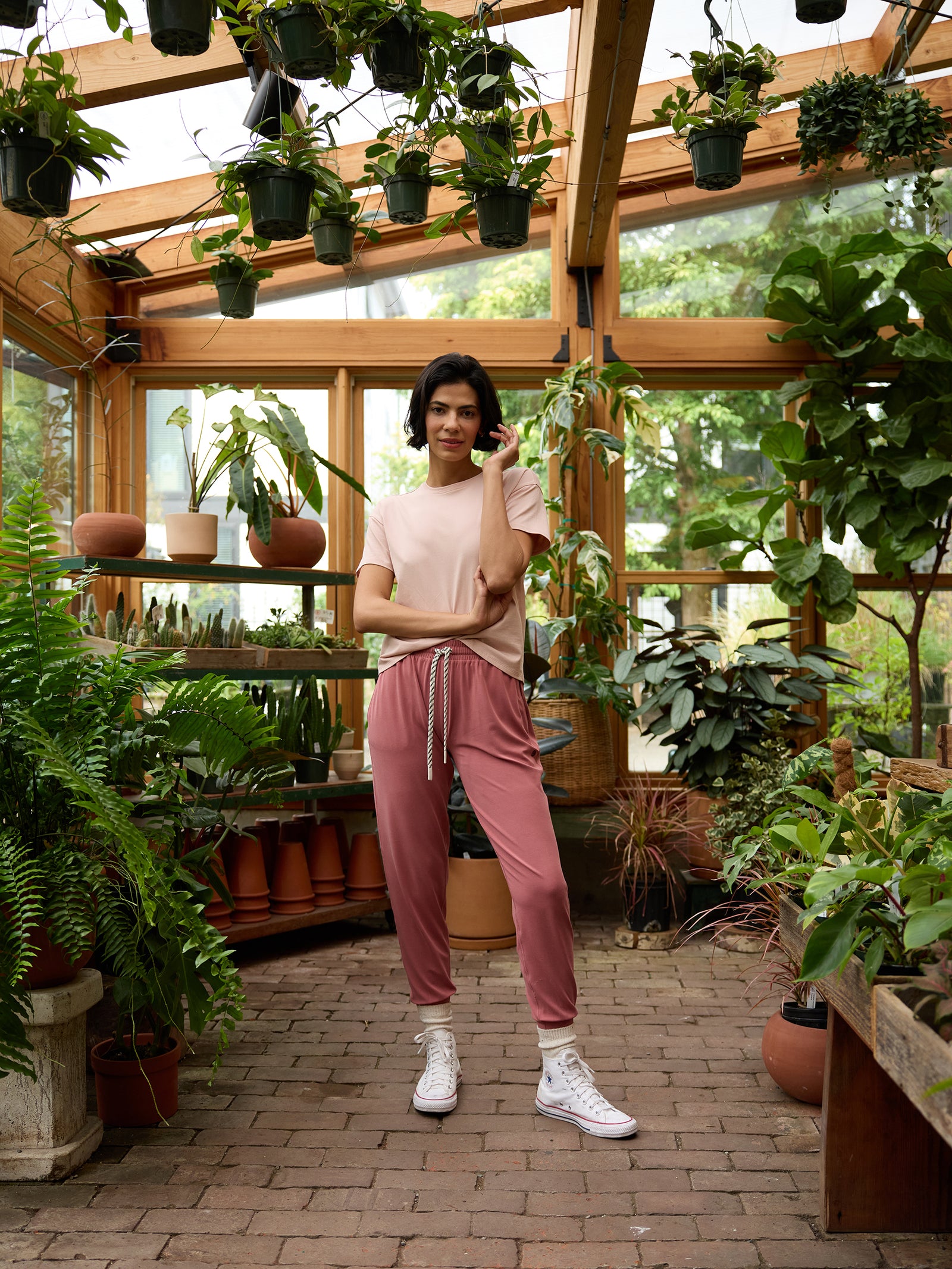 A person stands in a lush greenhouse filled with a variety of potted plants and hanging greenery. They are dressed in a light pink t-shirt, Cozy Earth's Women's Studio Jogger pants in mauve, and white sneakers. The person poses with one hand touching their face and the other on their hip, looking at the camera. 