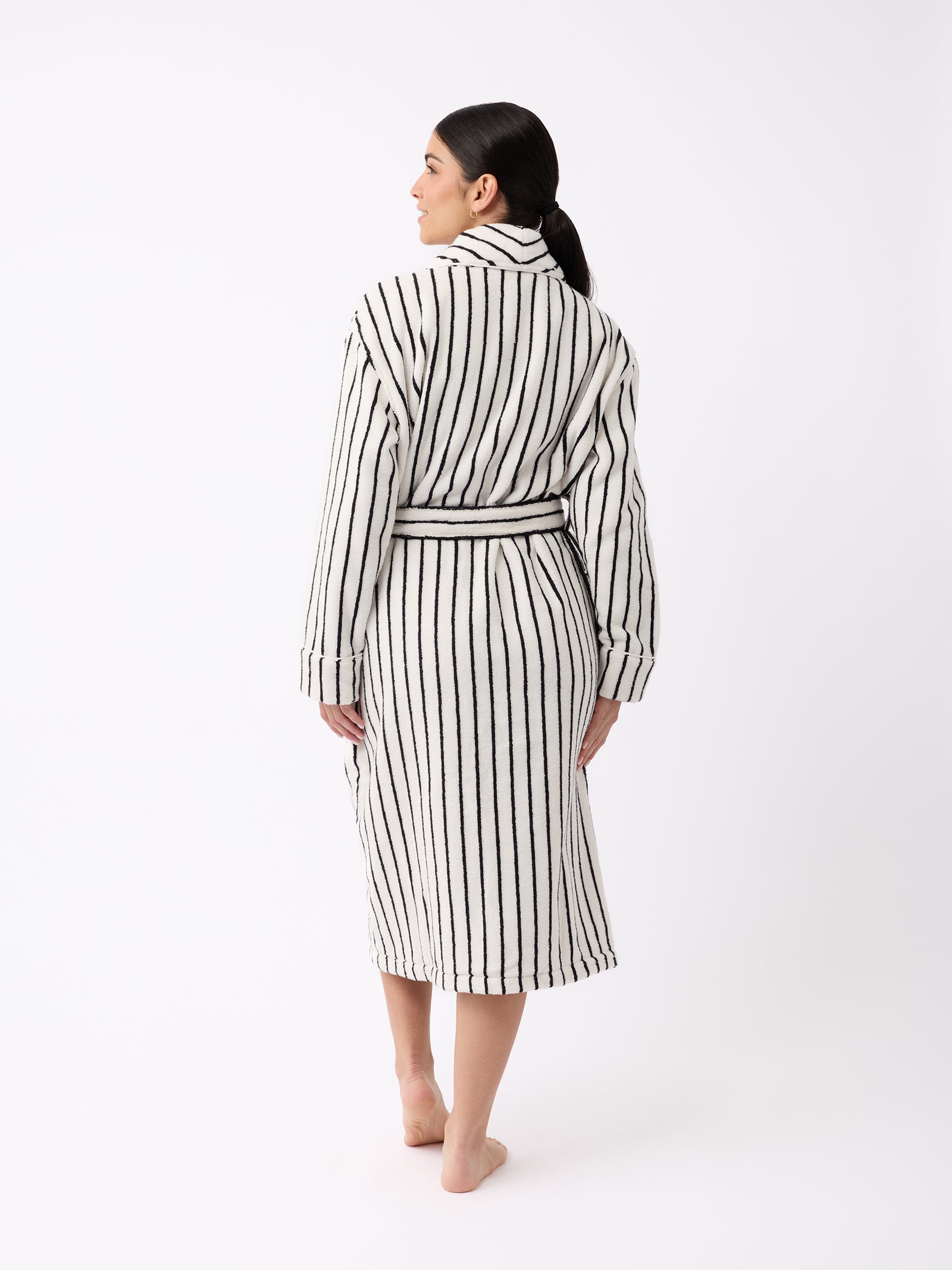 A person with dark hair stands barefoot on a white background, draped in the Luxe Bath Robe by Cozy Earth. The robe features black and white vertical stripes and is knee-length, cinched at the waist with a belt as they face away from the camera. 