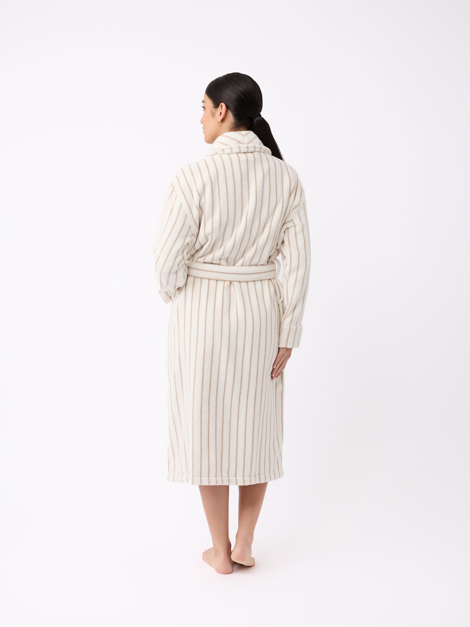 In a minimalist setting with a plain white background, an individual with long dark hair stands barefoot and faces away, adorned in Cozy Earth's Luxe Bath Robe. The robe features vertical beige stripes on a cream-colored fabric and is elegantly fastened at the waist with a belt. 