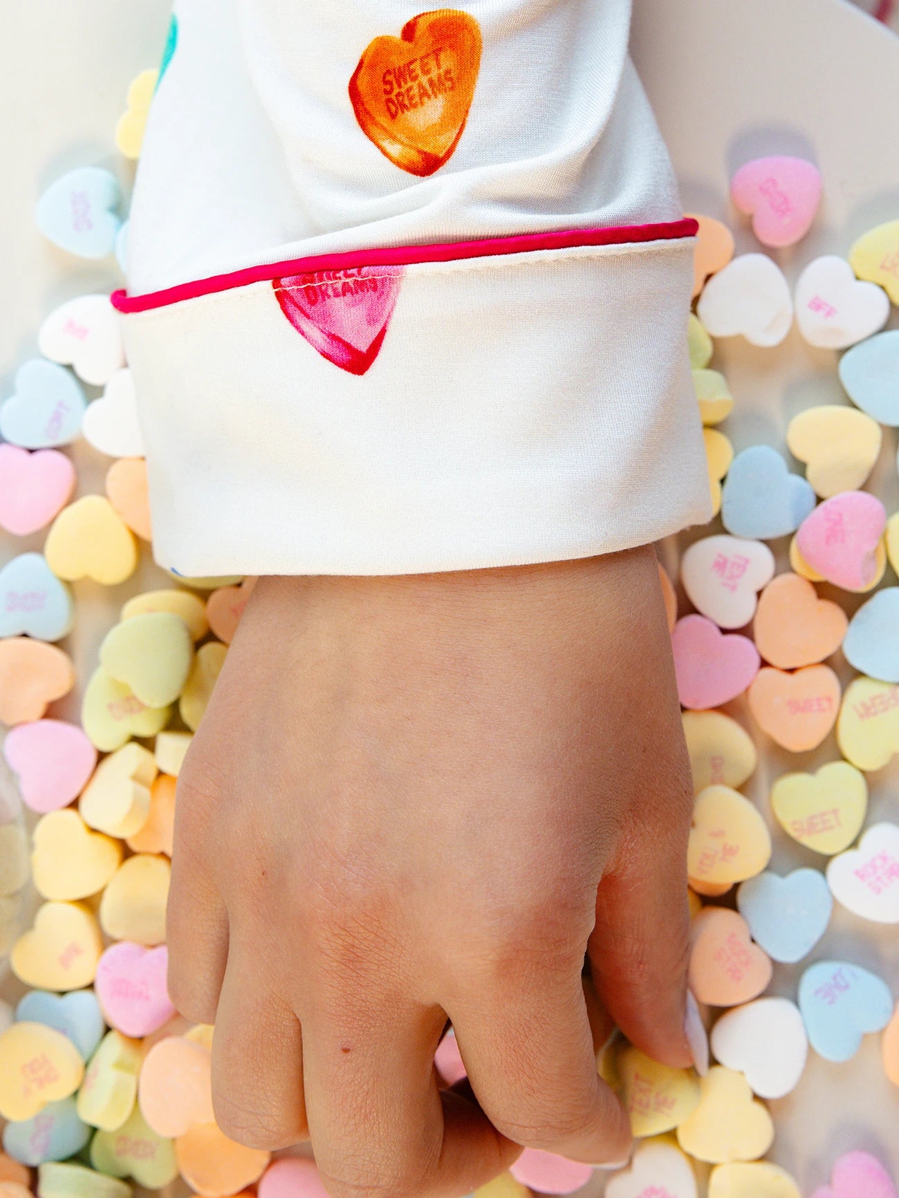 A hand rests on a pile of colorful, heart-shaped candy with messages like "Sweet Dreams." The person wears Cozy Earth Women's Long Sleeve Bamboo Pajama Top in Stretch-Knit, a white garment with red piped edges and candy heart designs. 