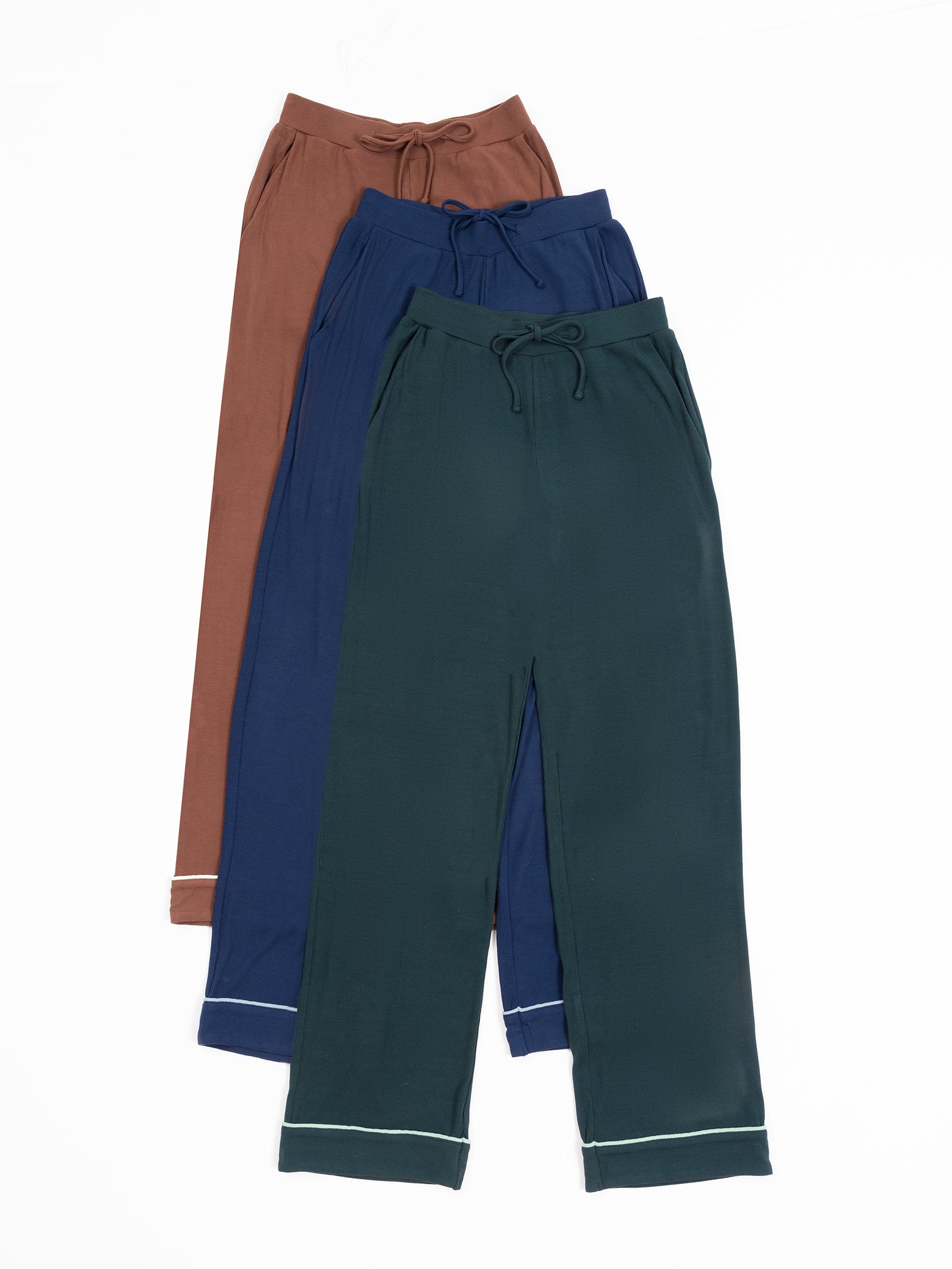 Three pairs of the Women's Bamboo Rib Knit Classic Pajama Pant by Cozy Earth are laid out on a white background. The pants are arranged in a stack with the bottom pair being dark green, the middle pair blue, and the top pair brown. Each pair features a drawstring waist and light-colored piping near the cuffs. 