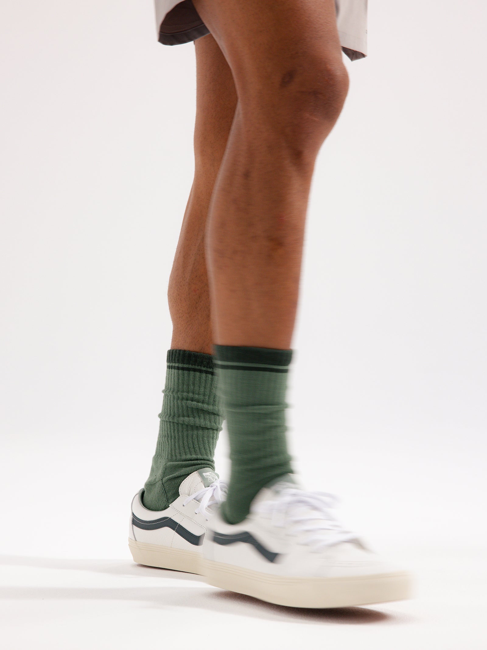 A person's lower legs are shown, wearing the Essential Calf Sock 2-Pack from Cozy Earth in green, paired with white sneakers featuring dark accents. The plain white background highlights the legs and footwear, capturing a mid-step motion. 