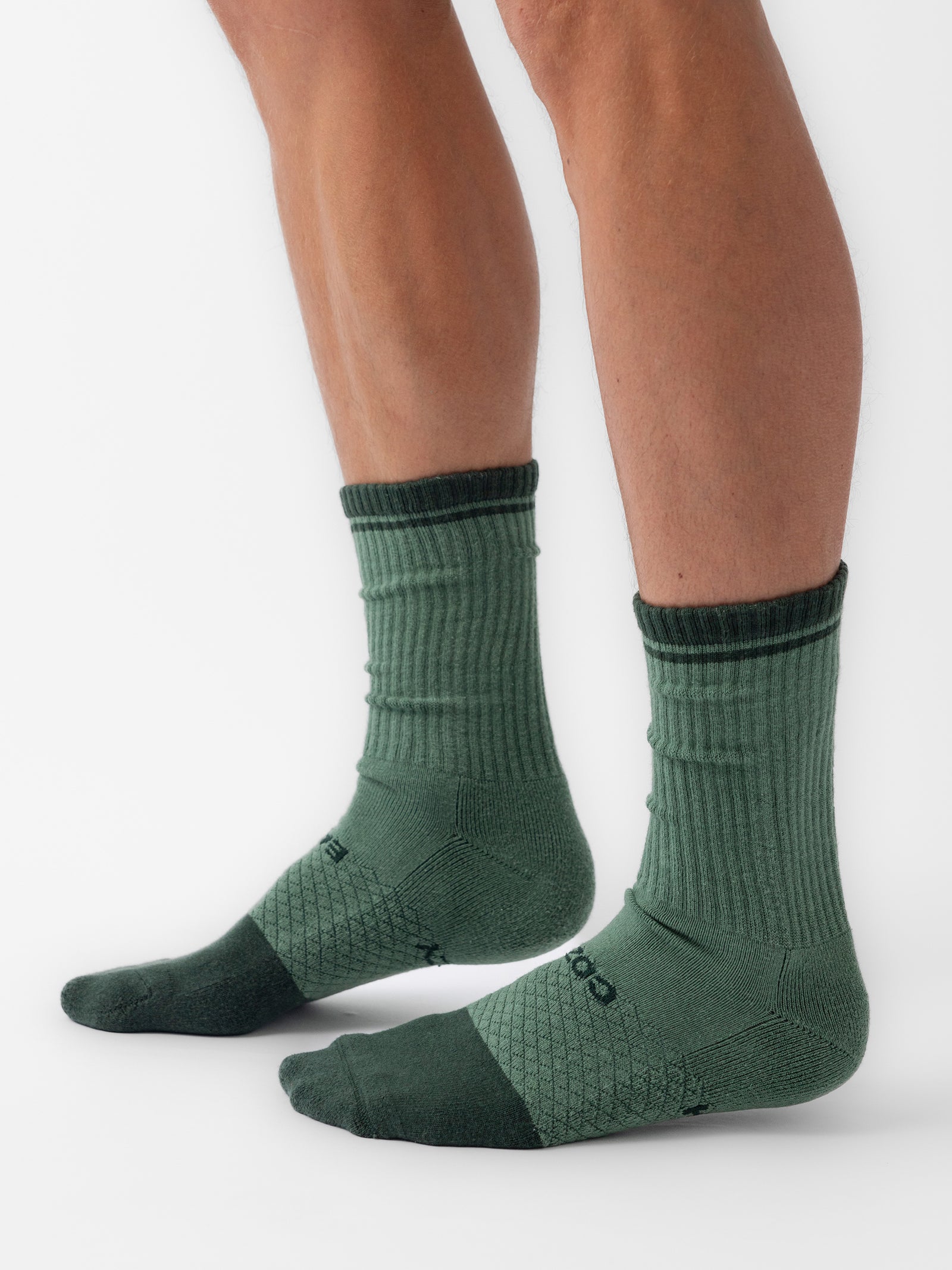 A person wearing Cozy Earth's Essential Calf Sock from the 4-Pack, showcasing a ribbed pattern on the calf and smoother fabric on the foot. The socks feature dark green toes and a dark green band around the upper edge. The person stands on a plain white background. 