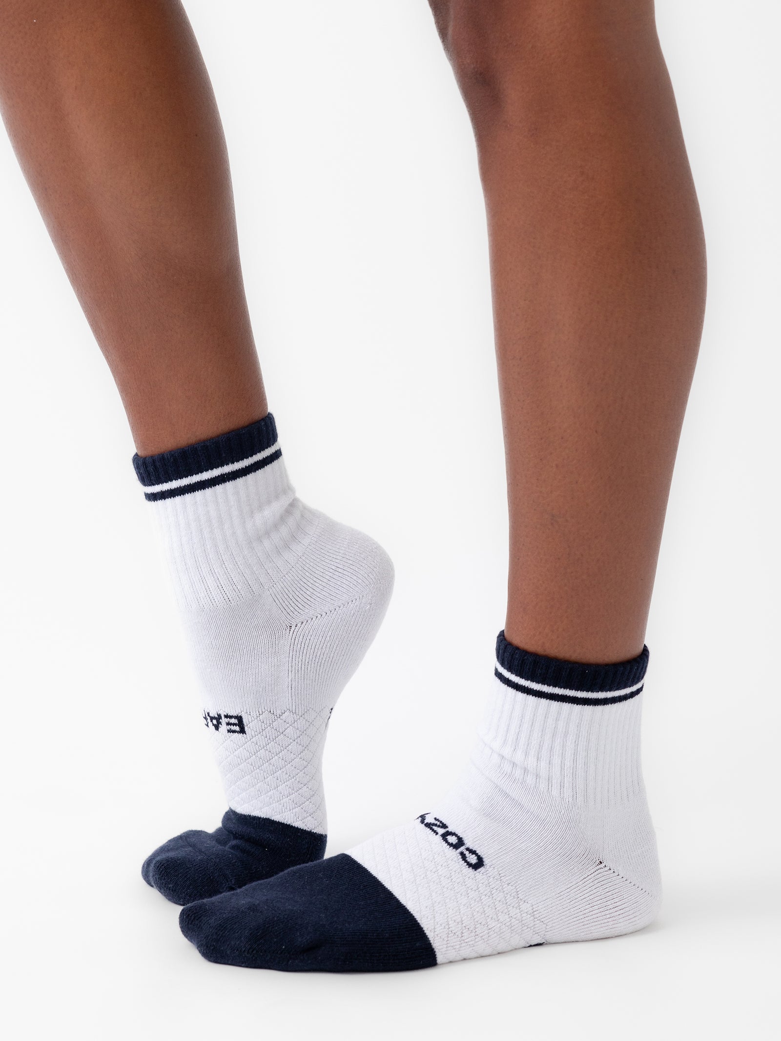 Close-up of a person's legs wearing white and navy blue ankle socks. The socks are branded with "Cozy Earth" and feature ribbed cuffs and textured soles. The photo is taken against a plain white background, highlighting the contrast between the socks and the skin tone from the Essential Quarter Sock 4-Pack collection. 