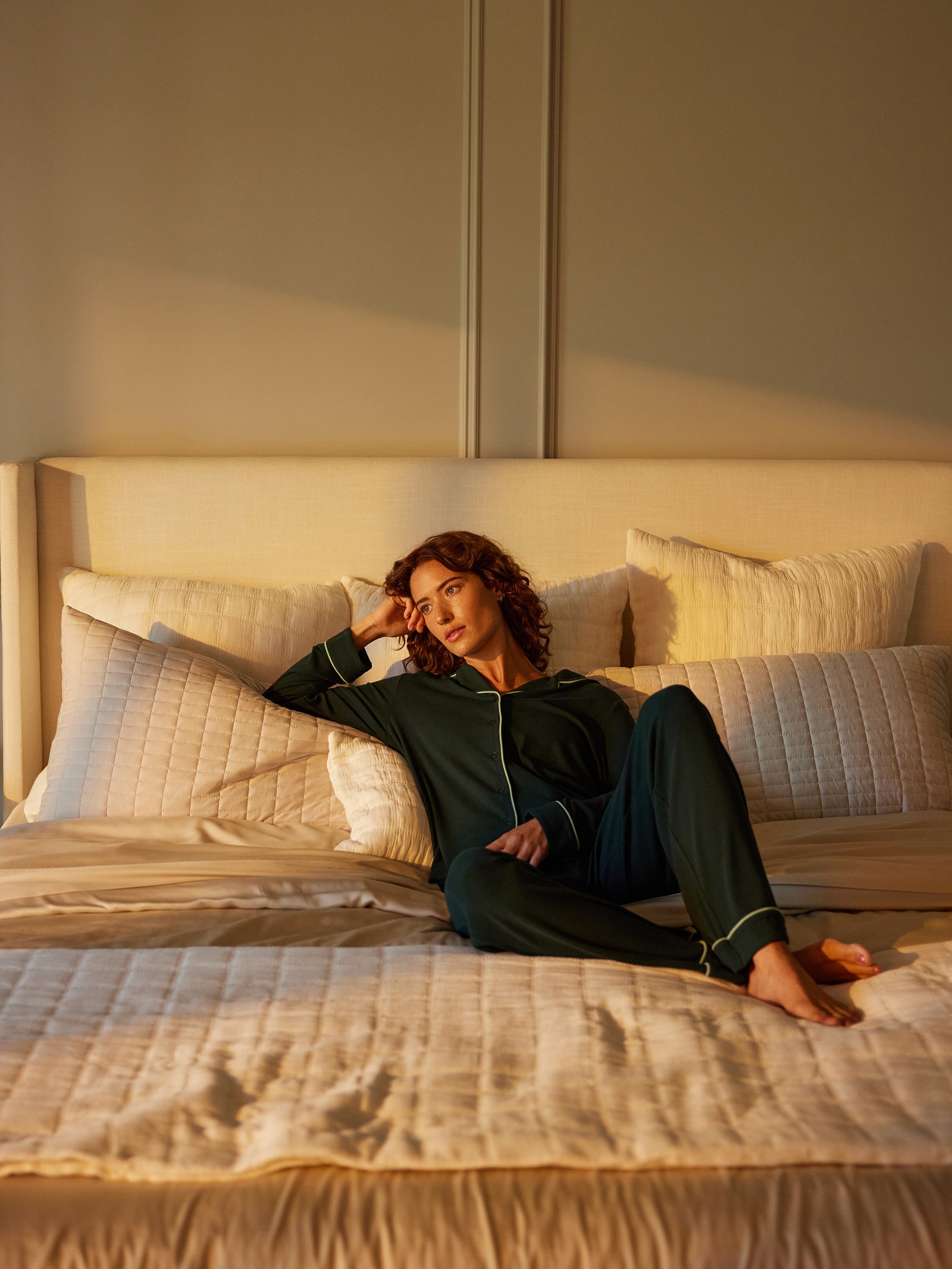 A person with curly hair is lounging on a beige bed while wearing Cozy Earth's dark green Women's Bamboo Rib Knit Classic Pajama Pant. They are resting their head on one hand, looking pensive. The room is softly lit by warm sunlight, and white and cream-colored pillows and bedding create a serene atmosphere. 