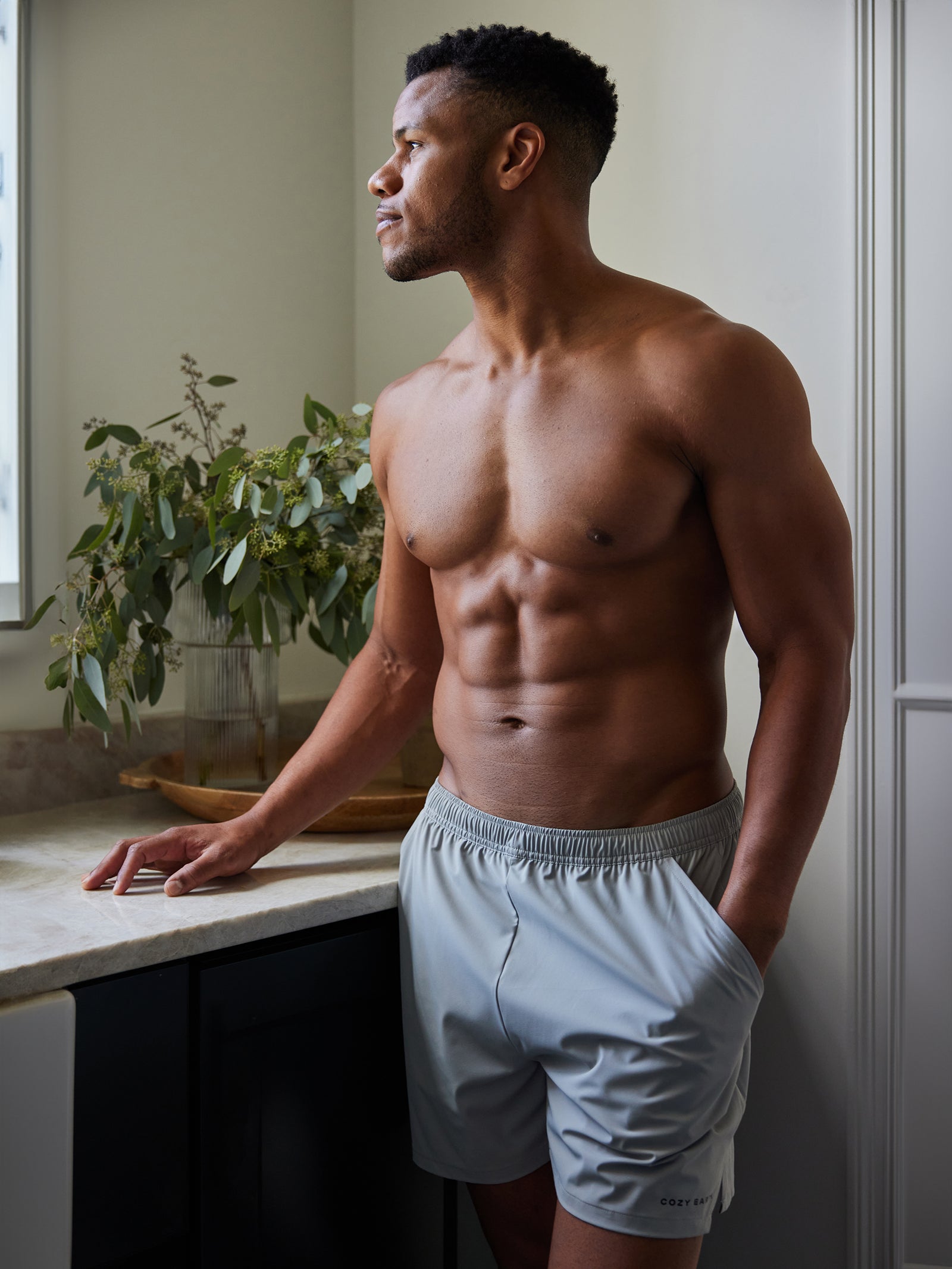 A shirtless man, wearing Cozy Earth's grey Men's Performance Sleep Short, stands in a bright and minimalistic bathroom. He leans against the counter, gazing out of the window, with a potted plant placed beside him. 