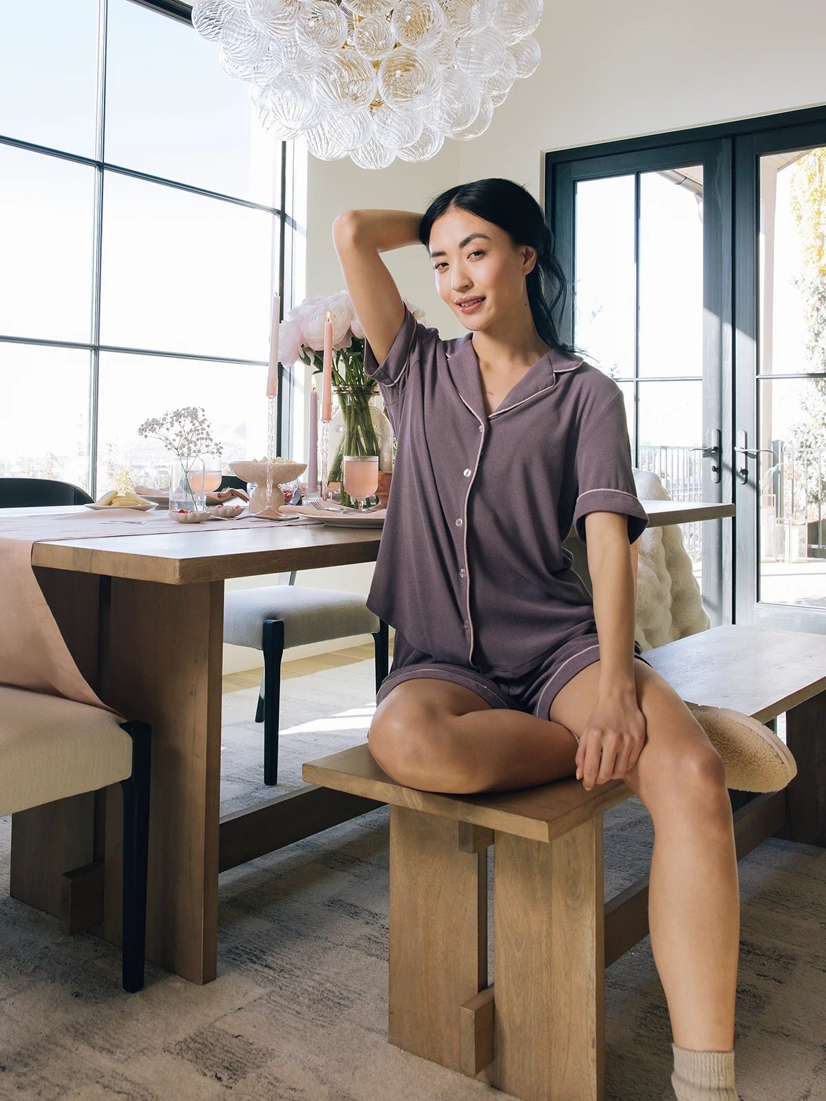 A woman in Cozy Earth's Women's Bamboo Rib-Knit Classic Pajama Short poses casually on a wooden bench at a dining table. The well-lit room has large windows and features a chandelier, elegantly set table with flowers, and tableware in the background. 