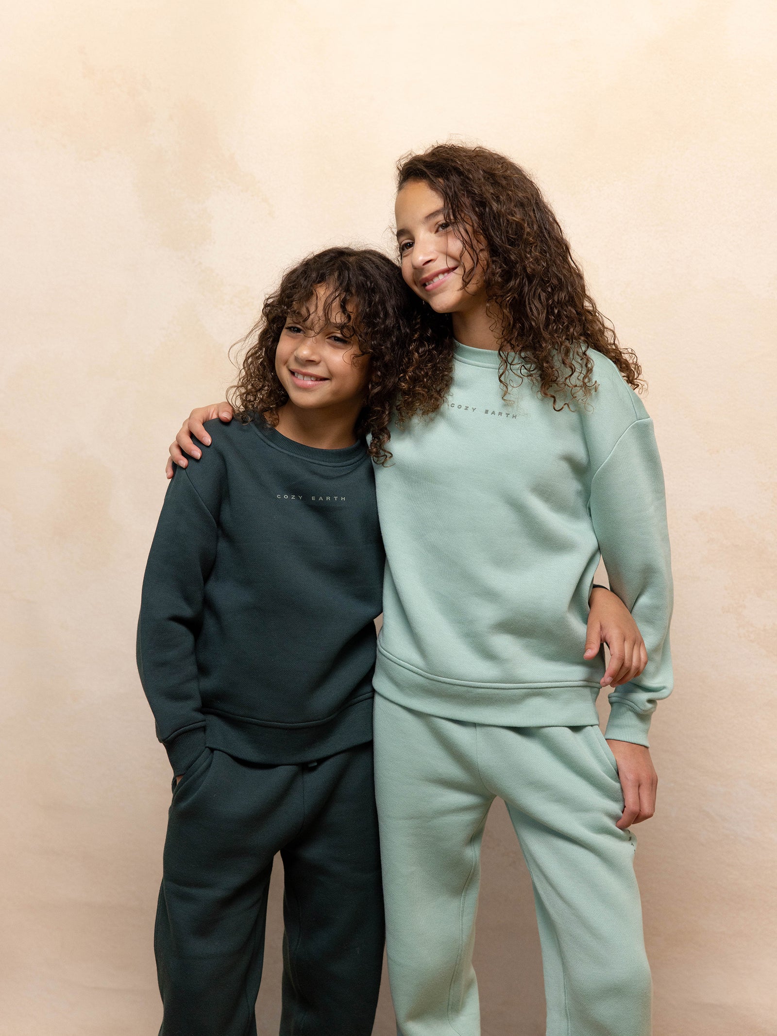 Two smiling children with curly hair are wearing Cozy Earth's Kid's CityScape Crewneck sweatshirts and pants. One is dressed in a dark green set while the other sports a light green ensemble. They pose affectionately against a neutral backdrop, with the older child wrapping an arm around the younger sibling. 