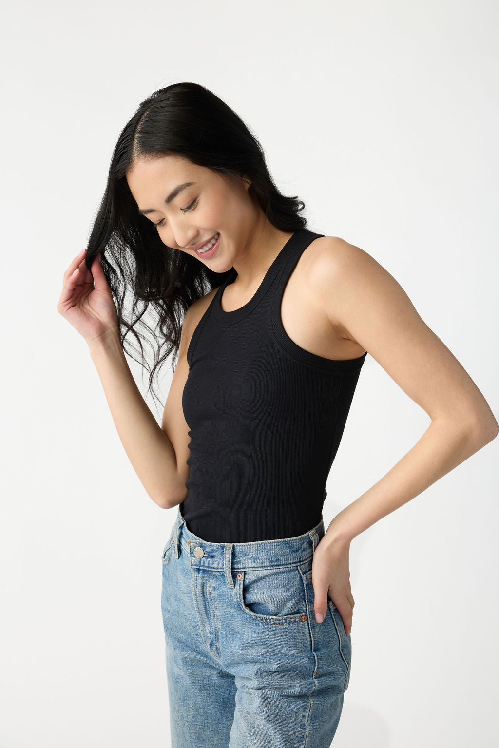 A dark haired woman wears Cozy Earth Women's Fine Ribbed Tank in Jet Black. She holds a strand of her hair, looks down, and stands in front of a white background. 