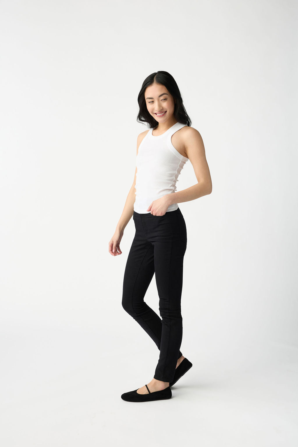 A dark haired woman wears Cozy Earth Women's Fine Ribbed Tank in White. She puts one hand in the pocket of her black jeans and stands in front of a white background. 