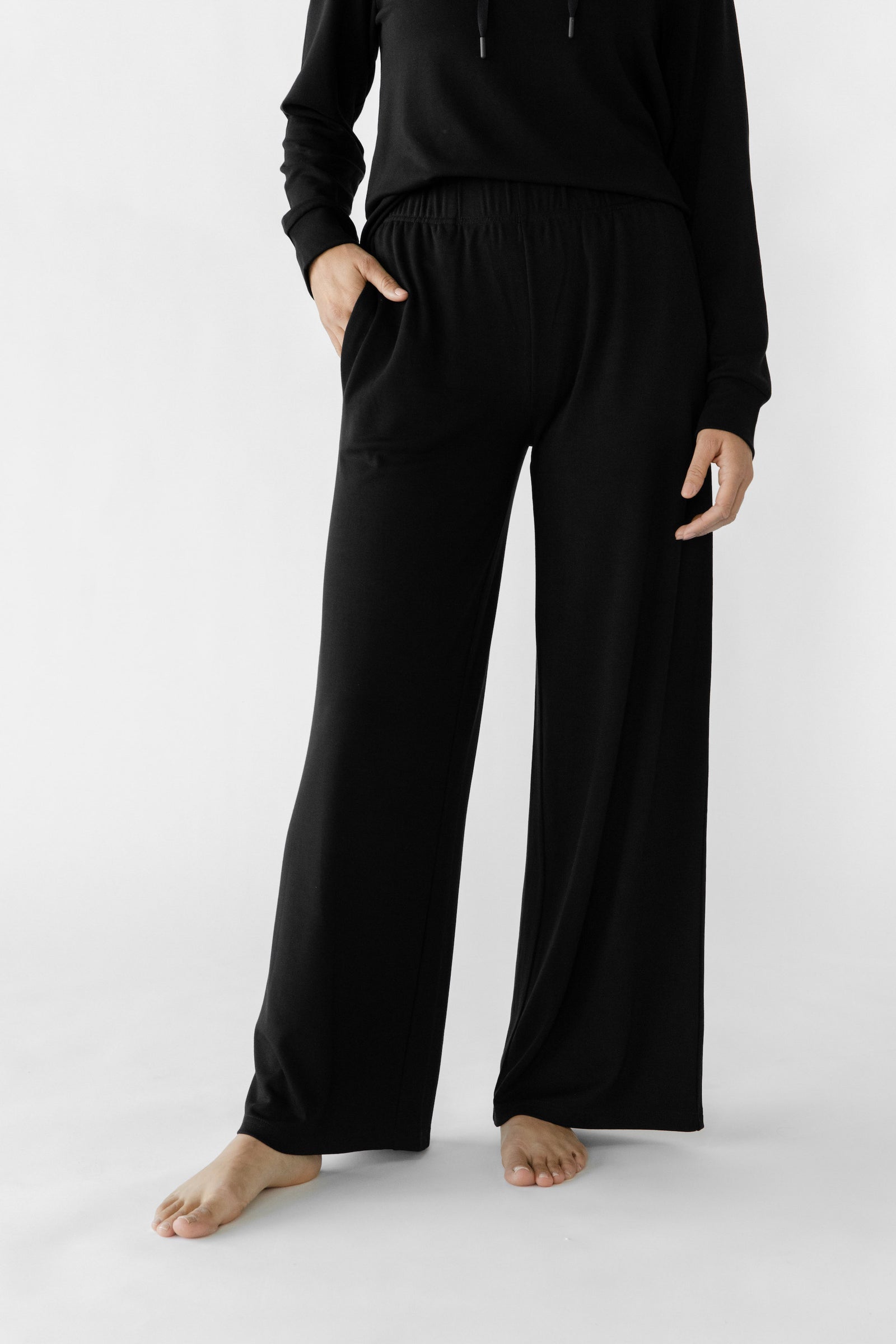 A person models the Women's Ultra-Soft Bamboo Wide Leg Pull On Pant & Hoodie Set by Cozy Earth, showcasing the black pants with pockets against a plain white background. The individual stands barefoot with hands resting in the pockets, head not visible. 