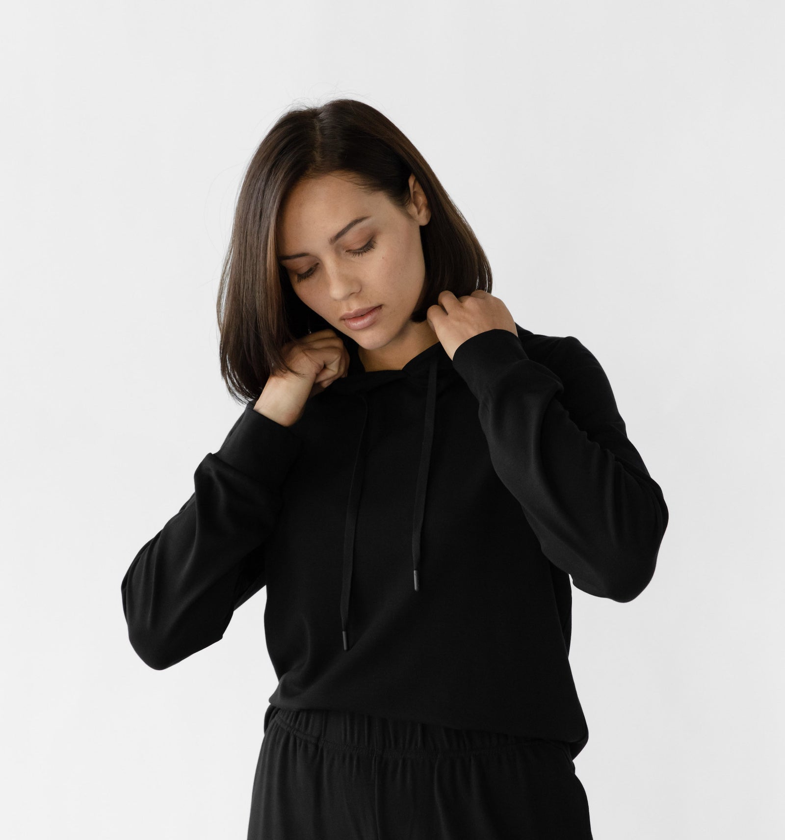 A woman with shoulder-length dark hair adjusts the hood of her black Cozy Earth Women's Ultra-Soft Bamboo Hoodie. She gazes downward and stands against a plain white background with a neutral expression. 