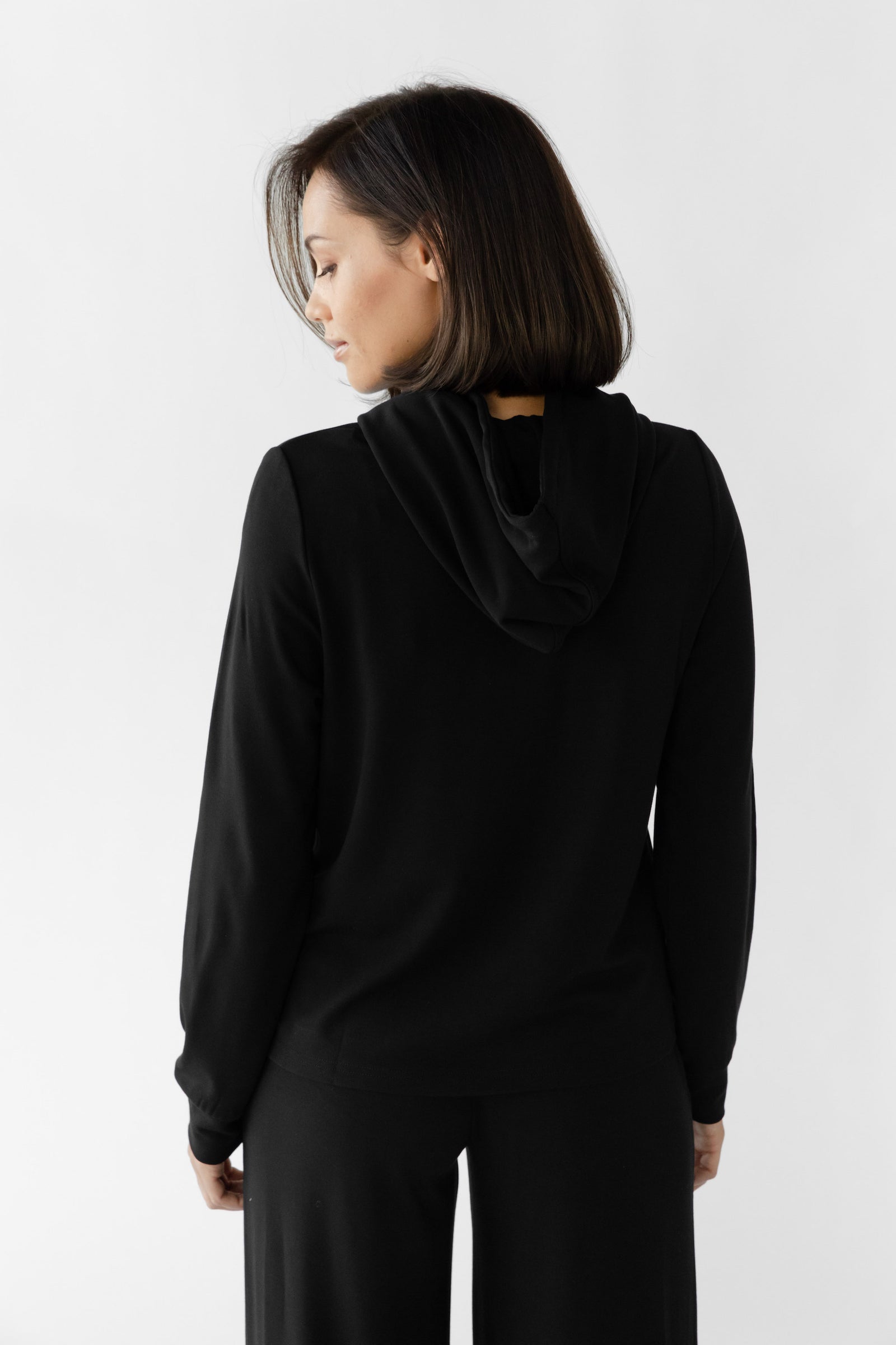 A person with shoulder-length hair stands back to the camera, slightly turned to the side, wearing Cozy Earth's Women's Ultra-Soft Bamboo Wide Leg Pull On Pant & Hoodie Set in black against a plain white background. 