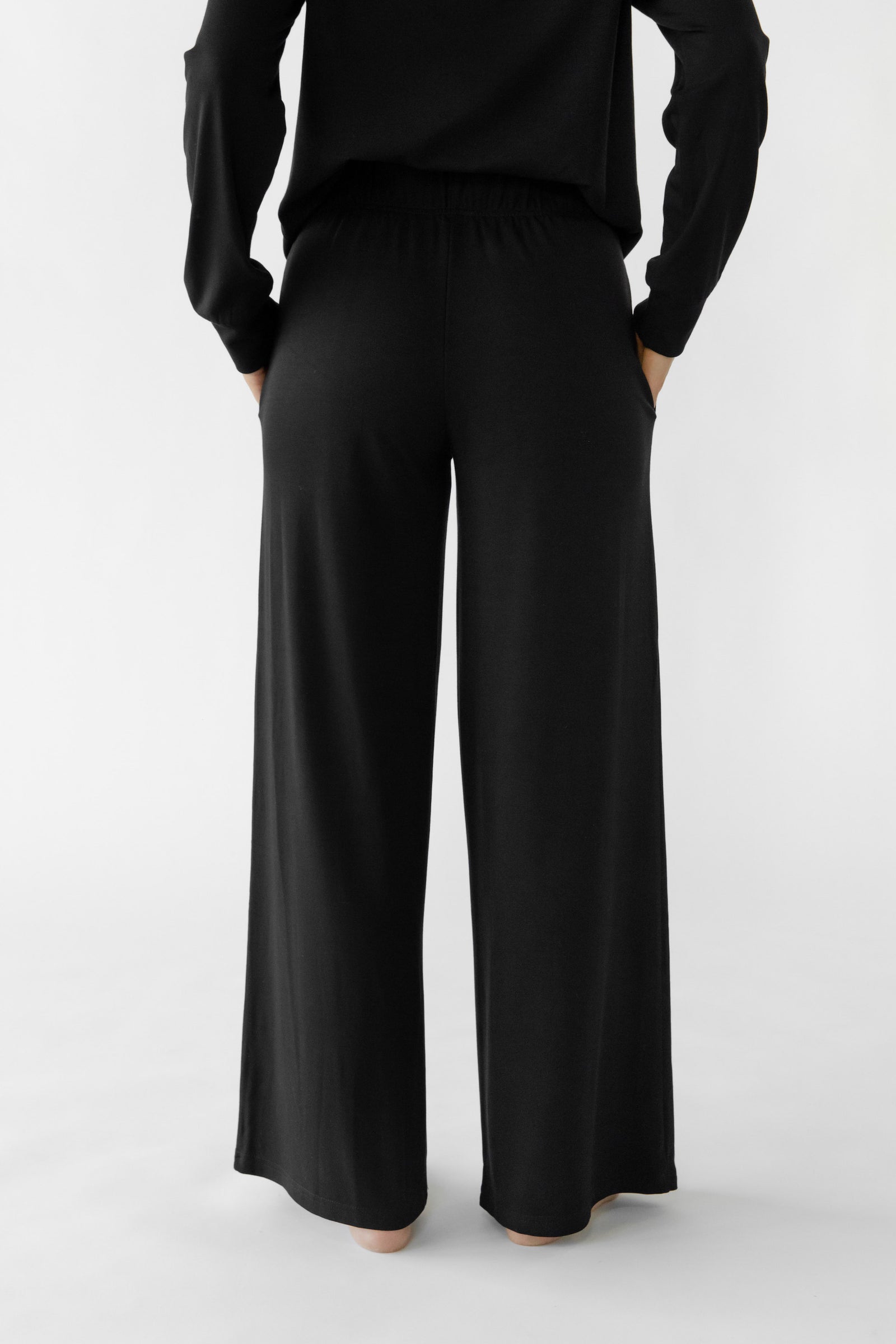 A person stands barefoot on a white background, wearing the Cozy Earth Women's Ultra-Soft Bamboo Wide Leg Pull On Pant & Hoodie Set in black, featuring loose-fitting pants with a wide leg and elastic waistband. 