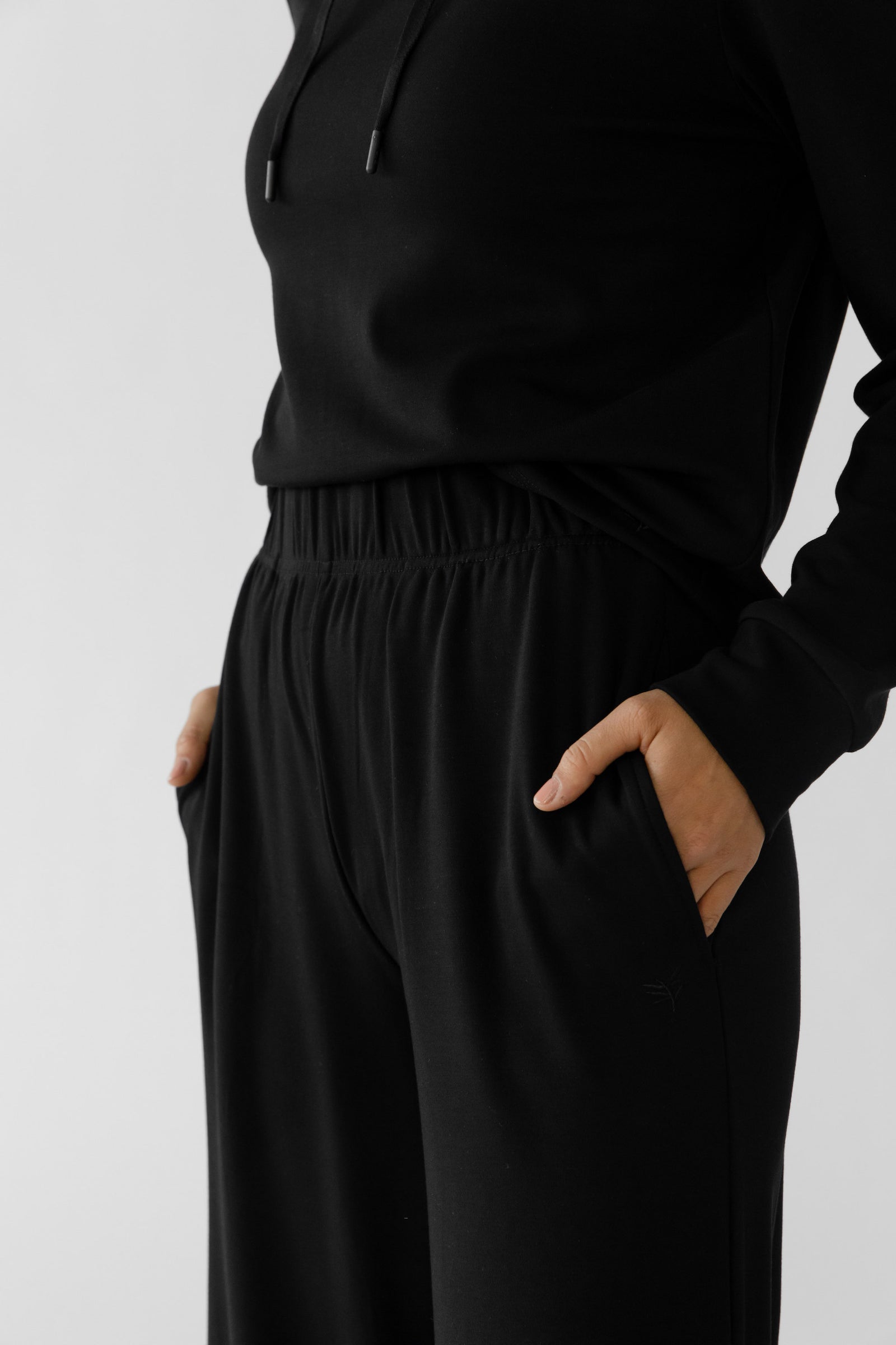 A person models the Cozy Earth Women's Ultra-Soft Bamboo Wide Leg Pull On Pant & Hoodie Set, showcasing the black hoodie and joggers against a plain white background, with hands casually in pockets. 